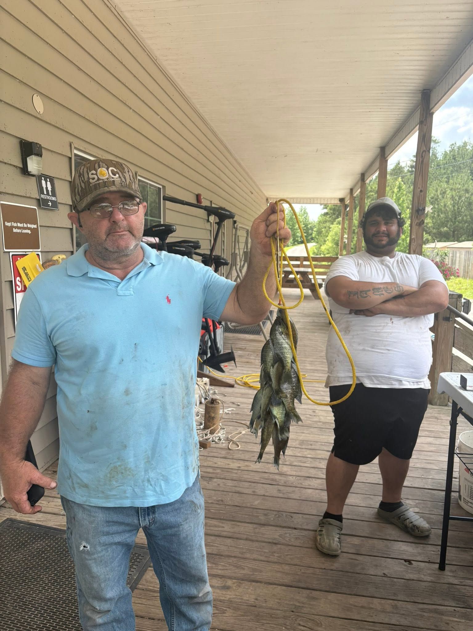 Two men on a porch holding a string of fish, one in a blue shirt and one in a white shirt.