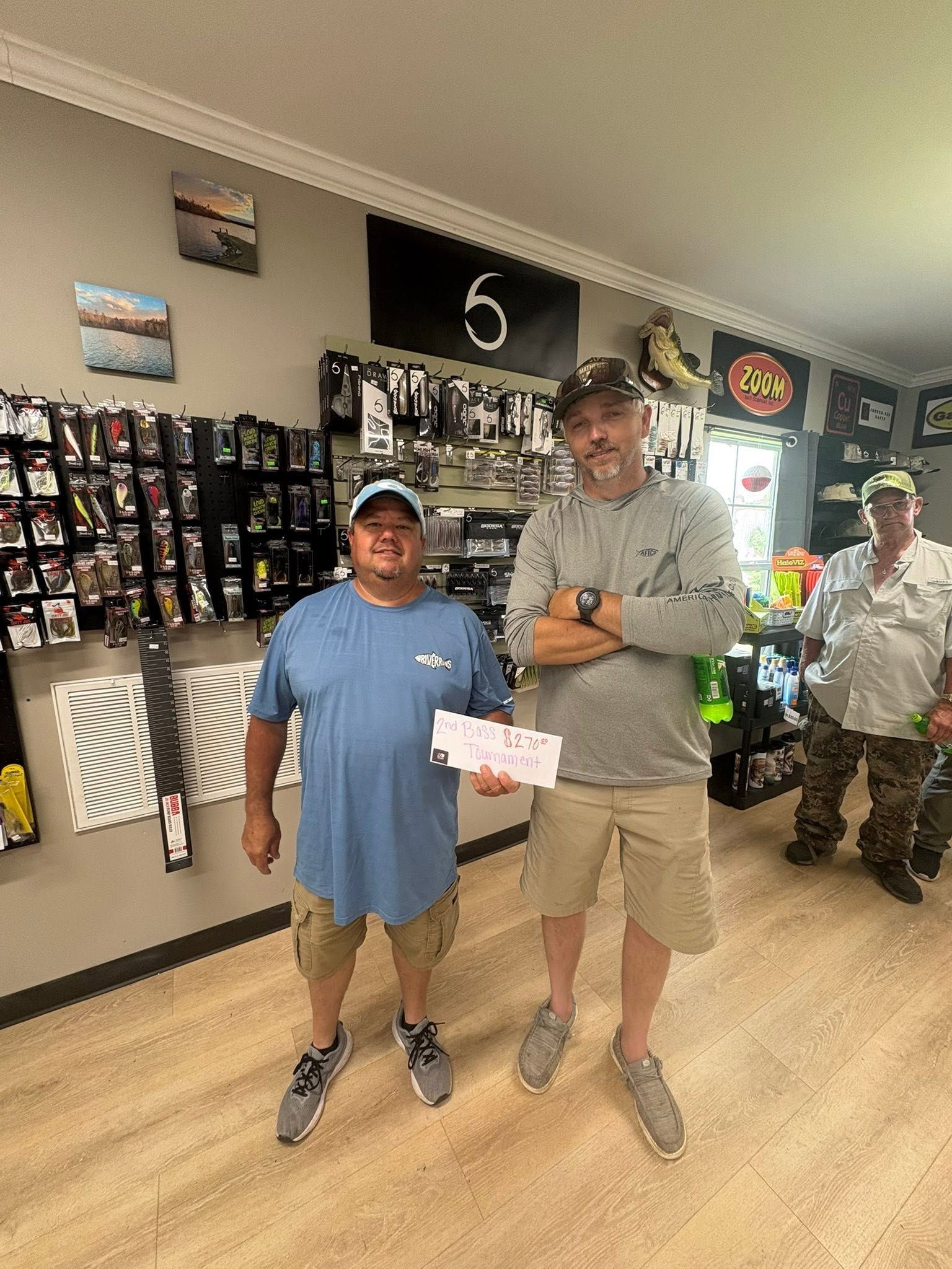 Two men stand inside a store, one holding a paper while the other holds a trophy.