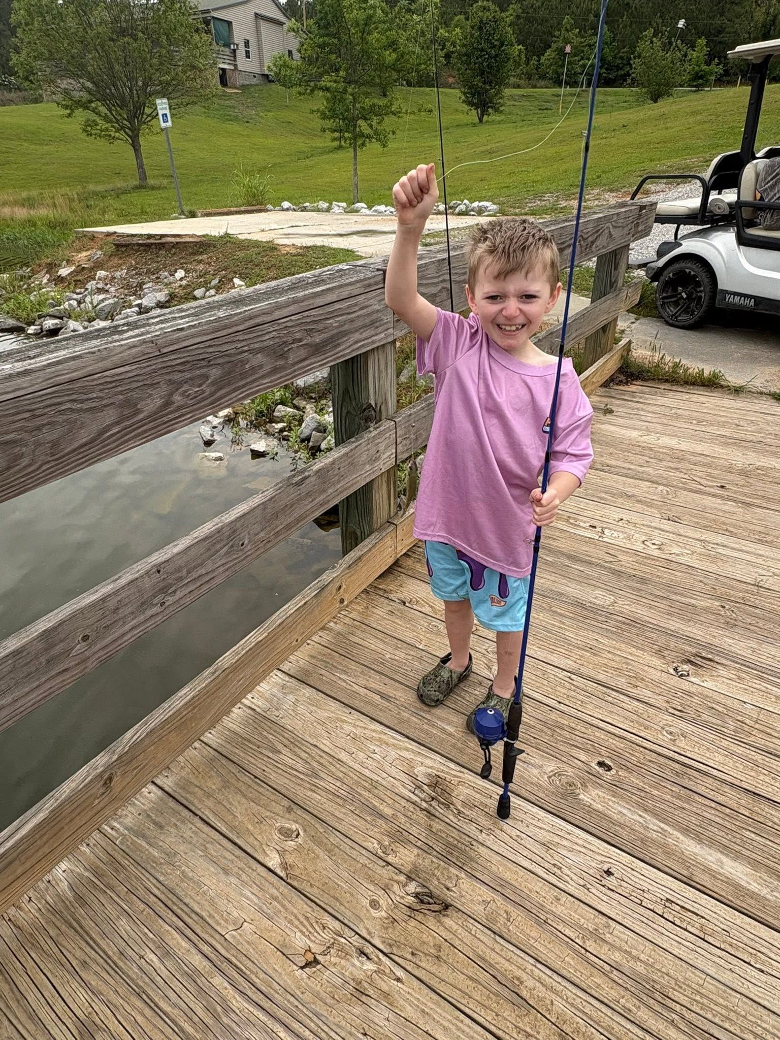 Child in a pink shirt standing on a wooden bridge, waving near a pond.