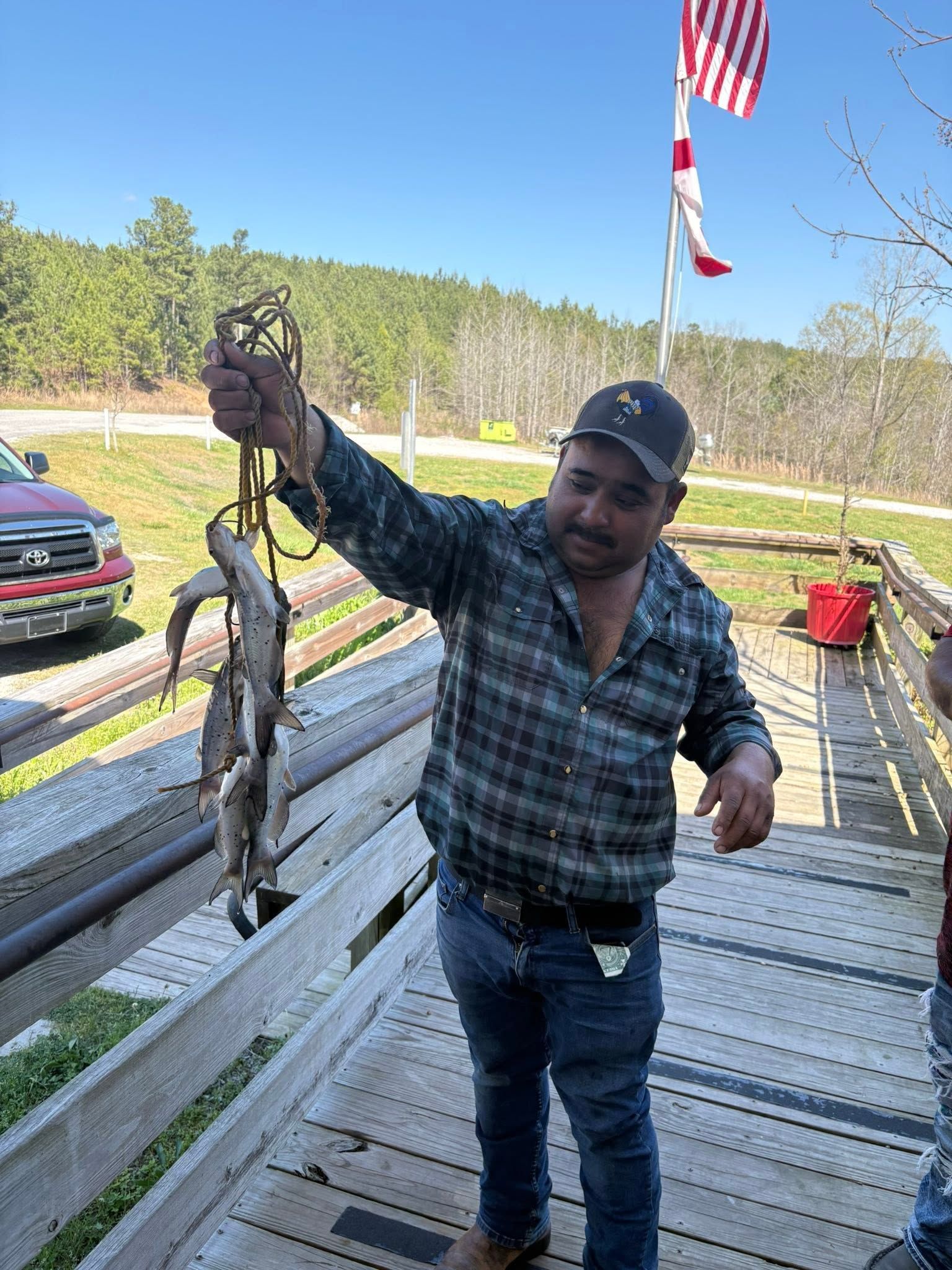 Man holding up a large fish on a wooden dock beside a parked truck and American flag