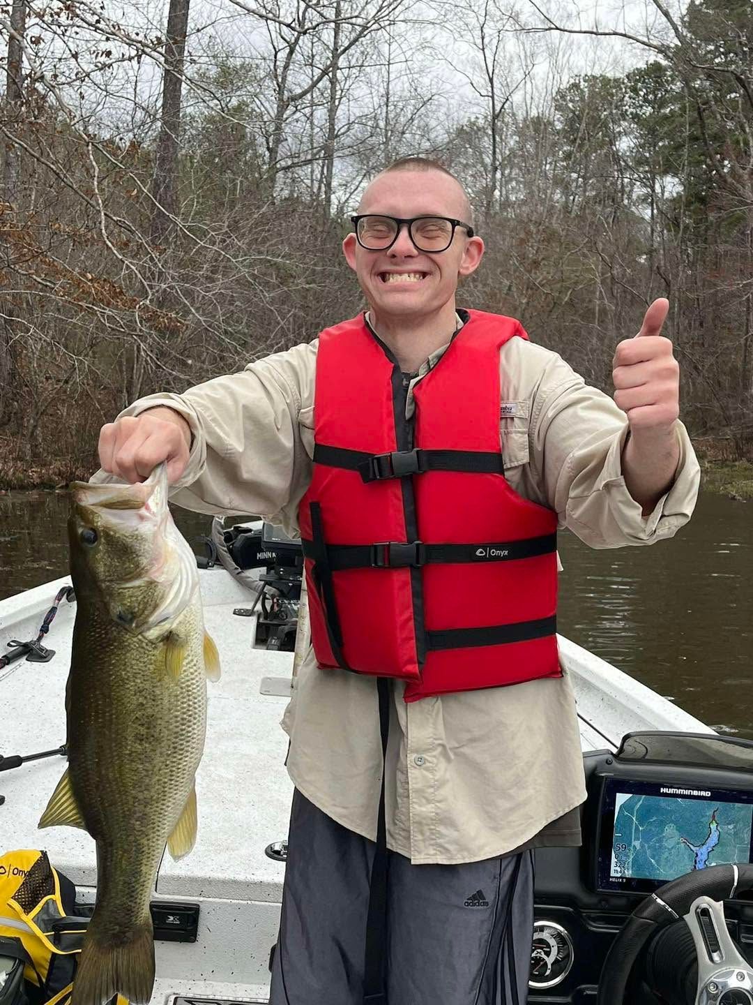 Smiling angler on a boat holding a large fish and giving a thumbs up in a wooded river setting