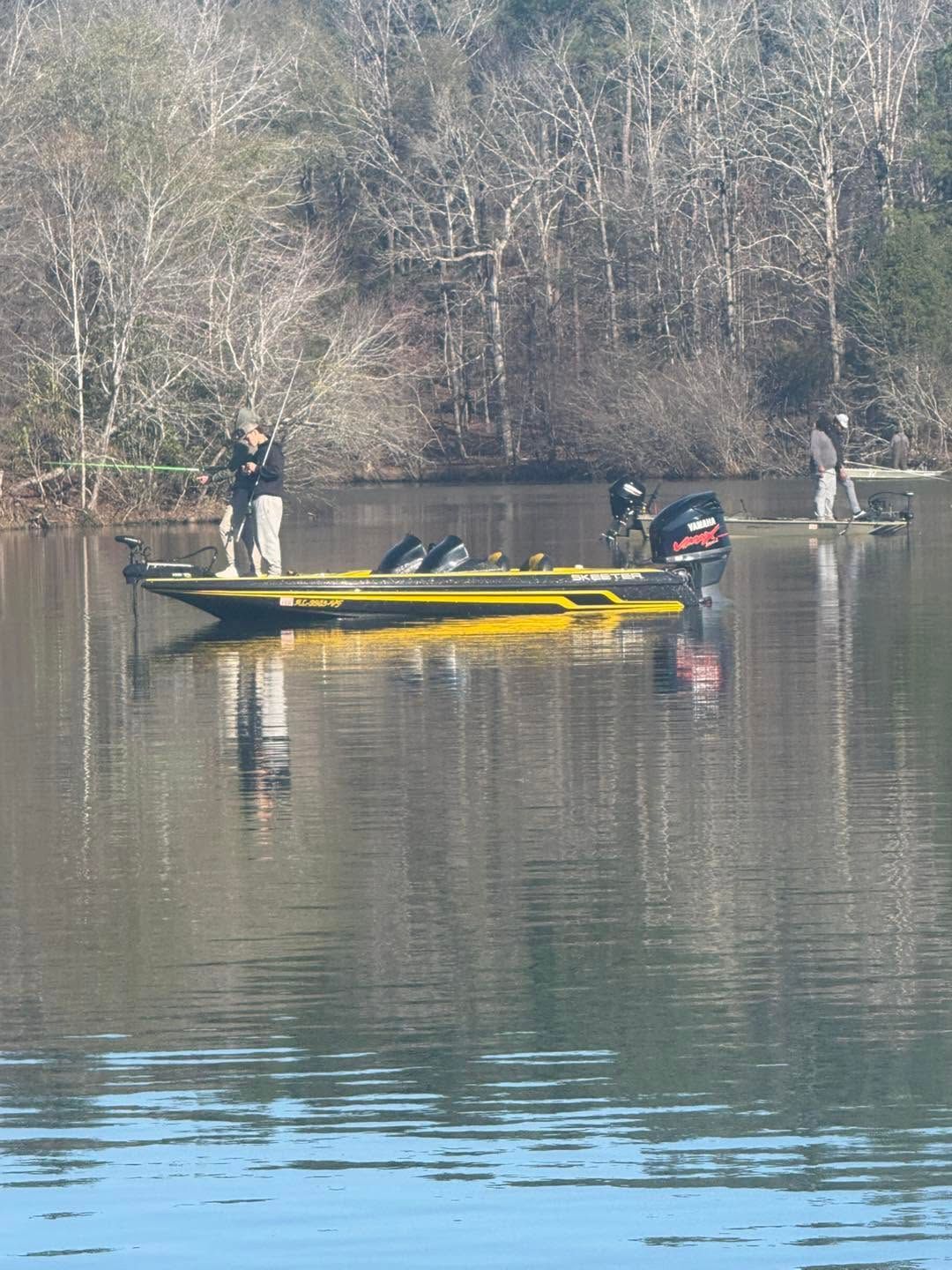 Two anglers on a yellow fishing boat on a calm lake, with bare trees reflected in the water.
