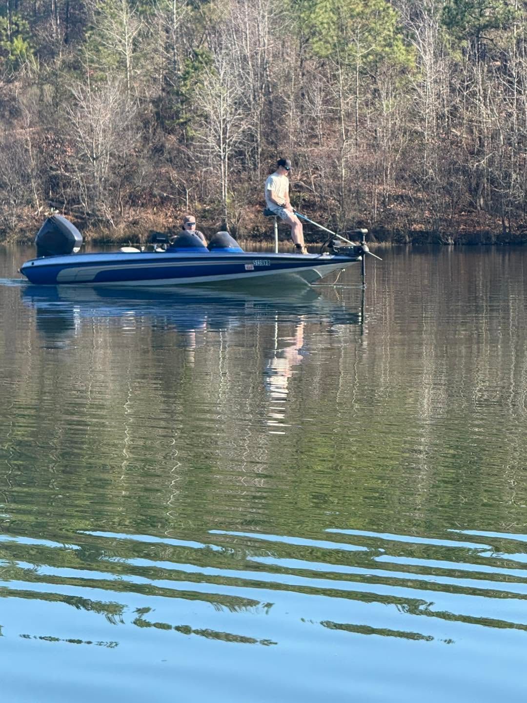 Fishing boat on a calm lake with two people, one standing and holding a rod, trees reflected on the water.