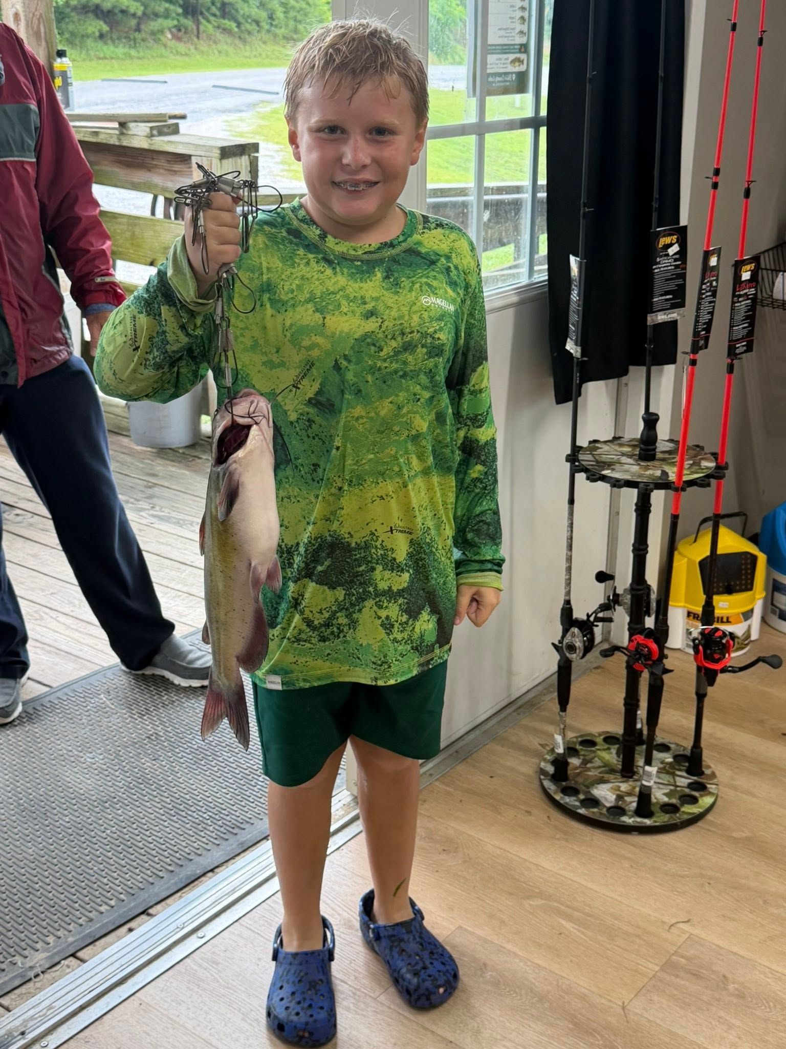 Boy in green shirt holding a large fish inside a shop by the doorway.