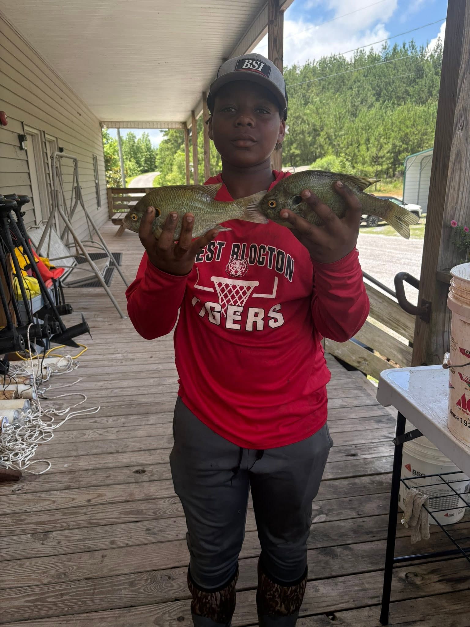 Person holding two large fish on a porch, wearing a red shirt and black cap.
