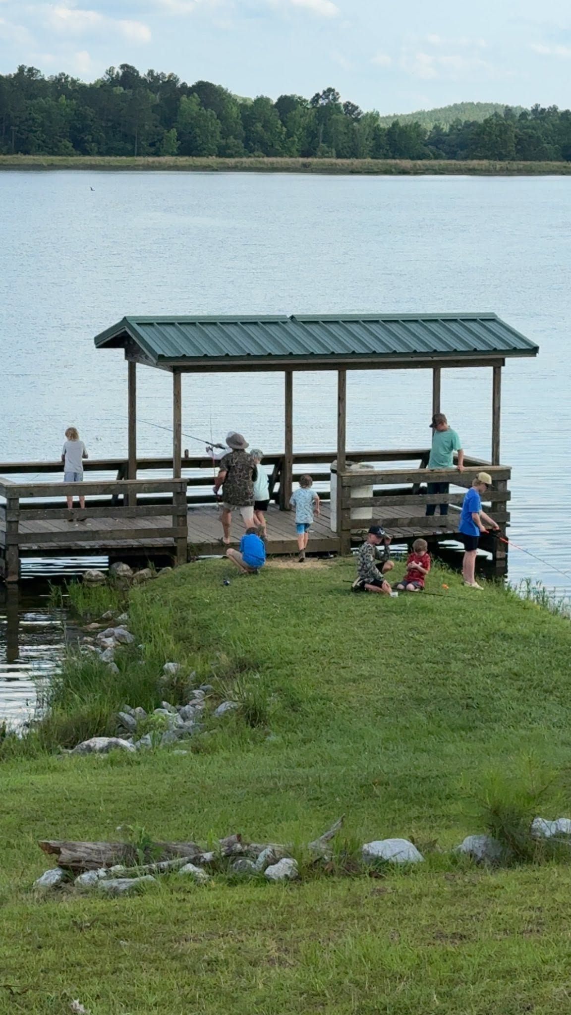 People under a small dock shelter by a calm lake, with trees across the water.