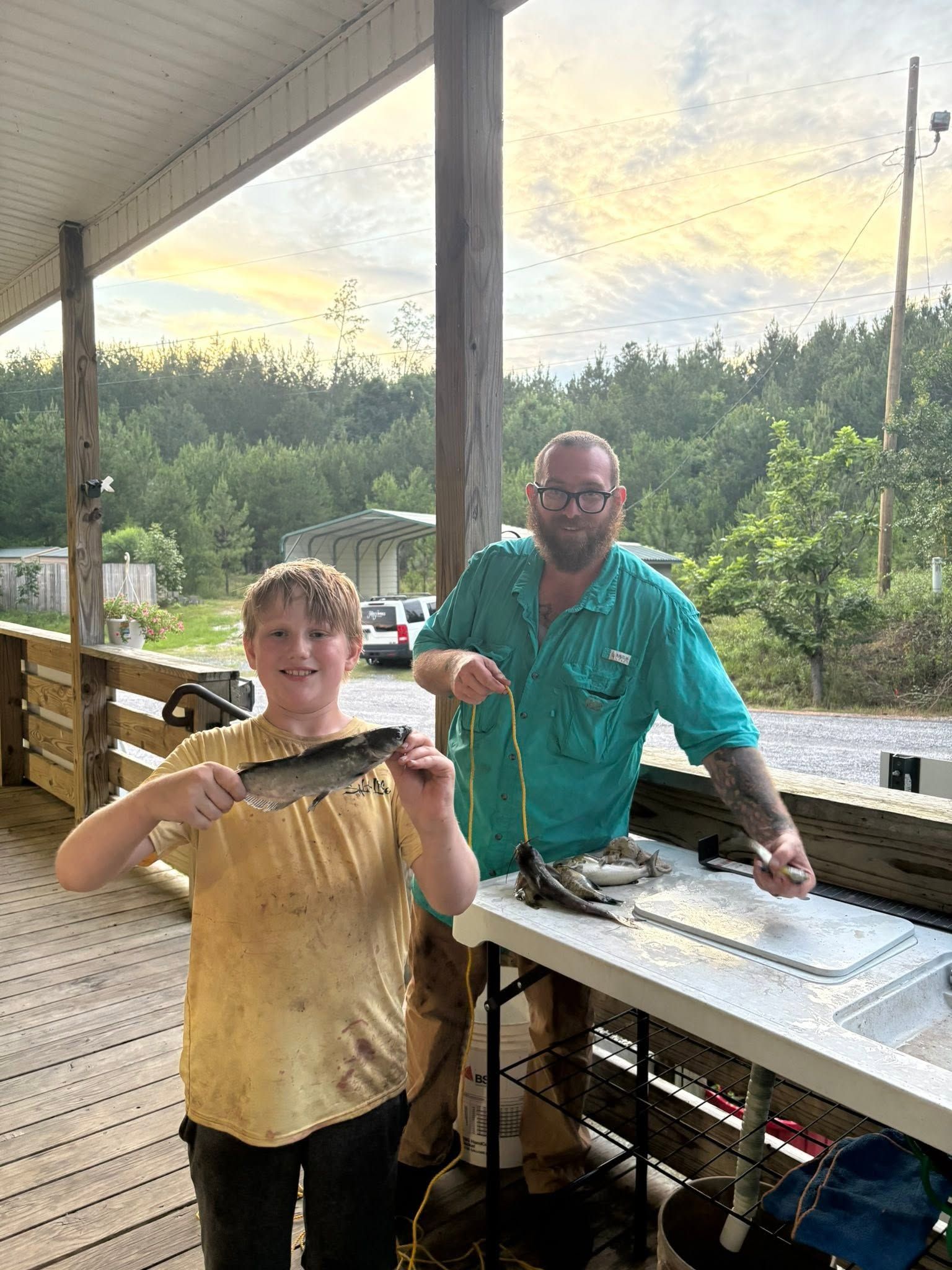 Two people on a porch holding a large fish and a knife, with a fish-cleaning table and pond in the background