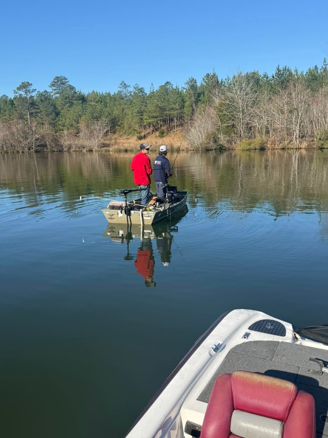 Two people standing on a small boat on calm water, with a red seat in the foreground and trees along the shore.