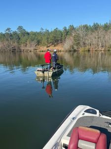 Two people standing on a small boat on calm water, with a red seat in the foreground and trees along the shore.