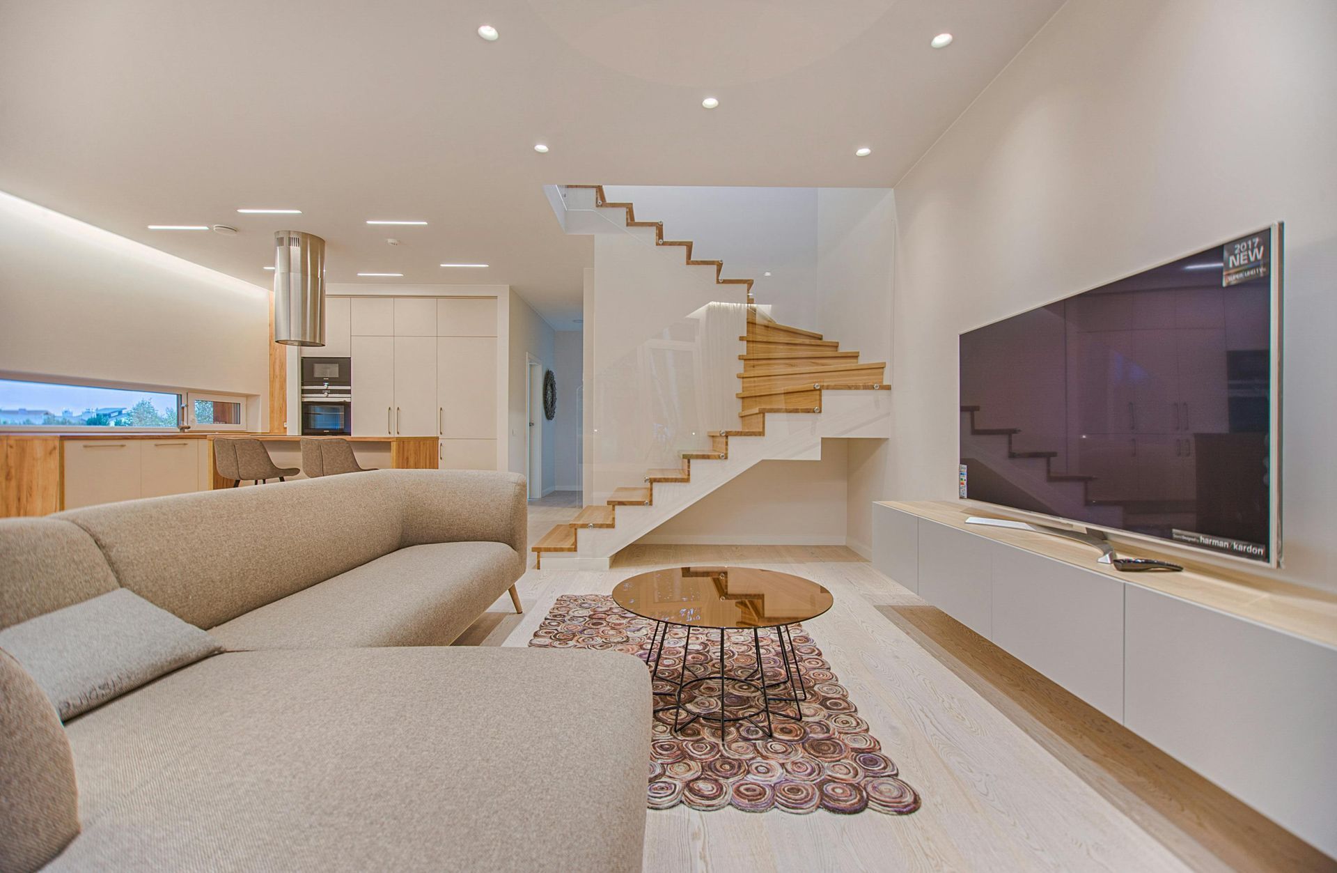 Modern living room with light-colored sofa, floating TV, wooden staircase, and open kitchen.