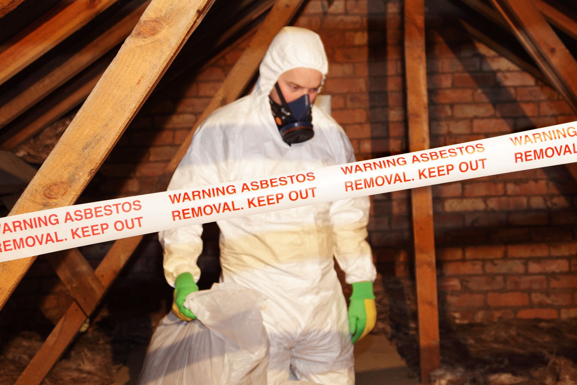 Person in protective suit removing asbestos from an attic. Warning tape present.