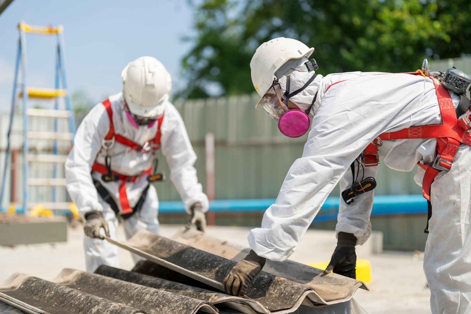 Two workers in protective suits removing asbestos roofing materials outdoors.