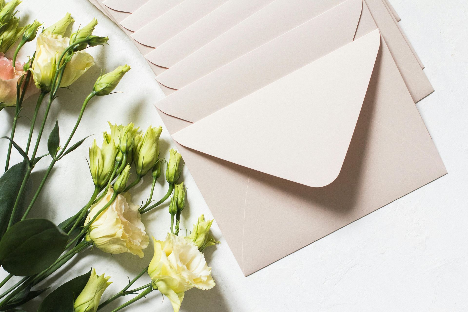 A stack of pale pink envelopes resting next to a bouquet of cream-colored Eustoma flowers on a white surface.