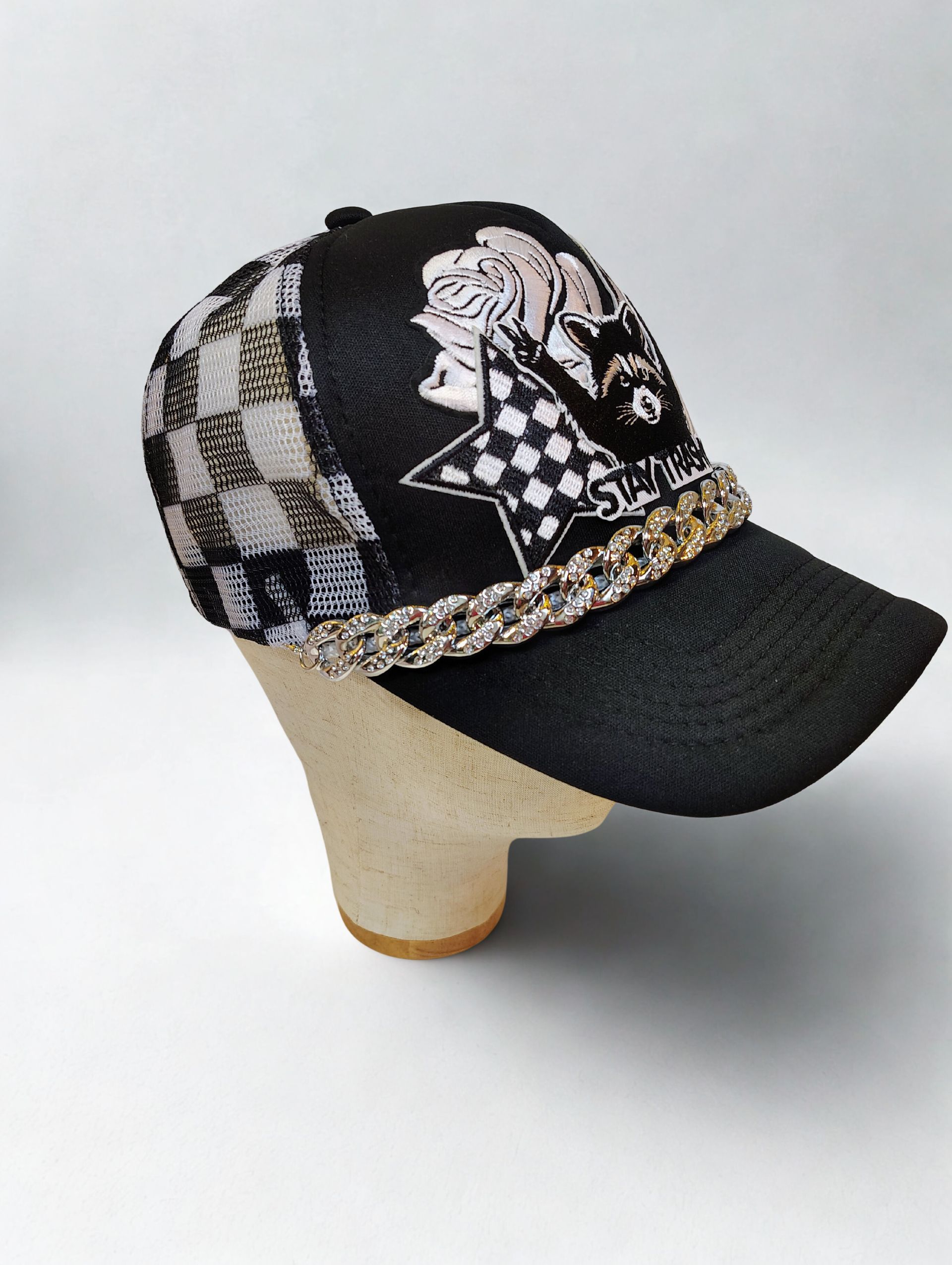 A black textured baseball cap featuring a raccoon graphic, a white star, checkered hearts, and a gold rhinestone chain.