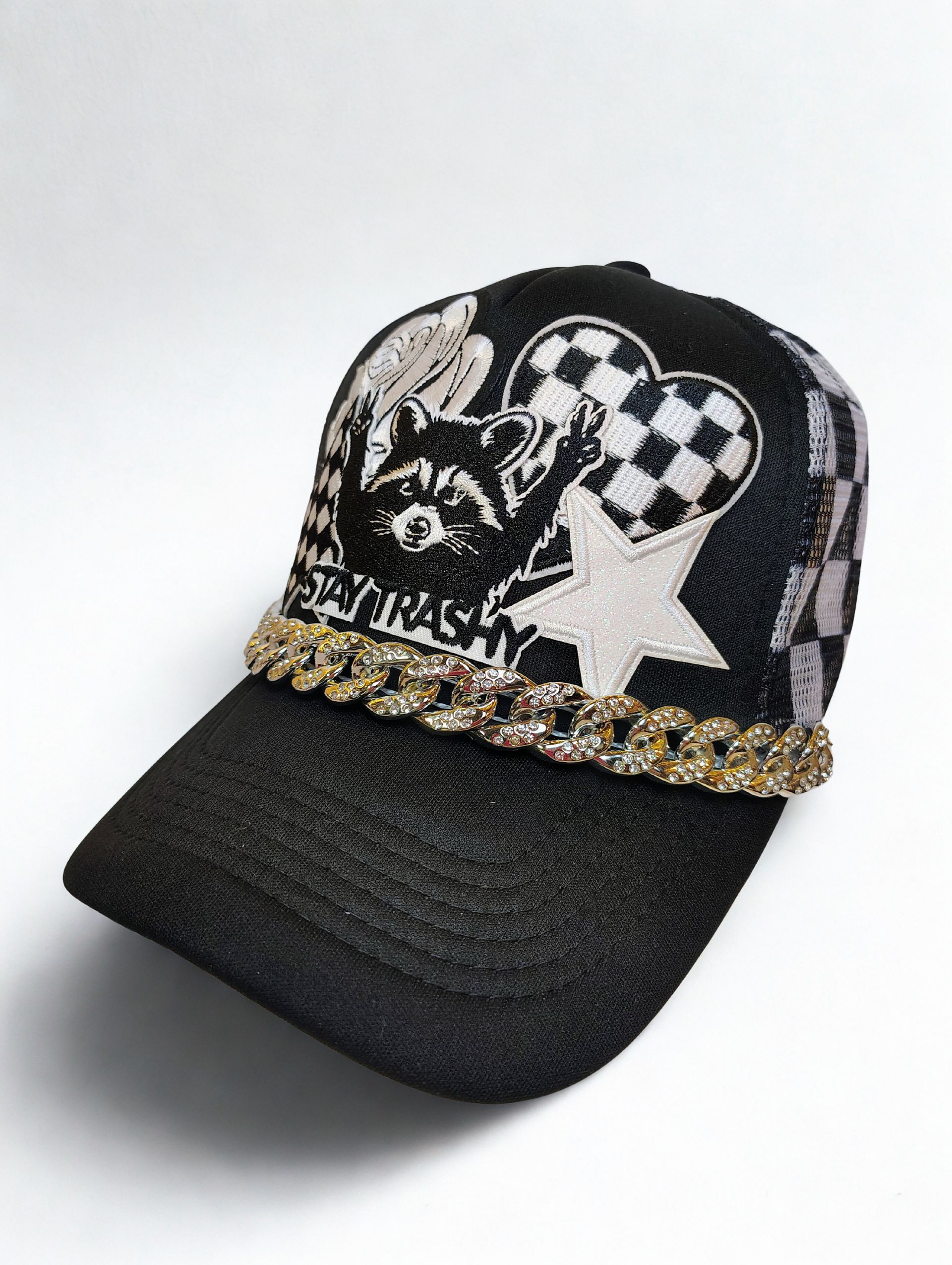 A black textured baseball cap featuring a raccoon graphic, a white star, checkered hearts, and a gold rhinestone chain.