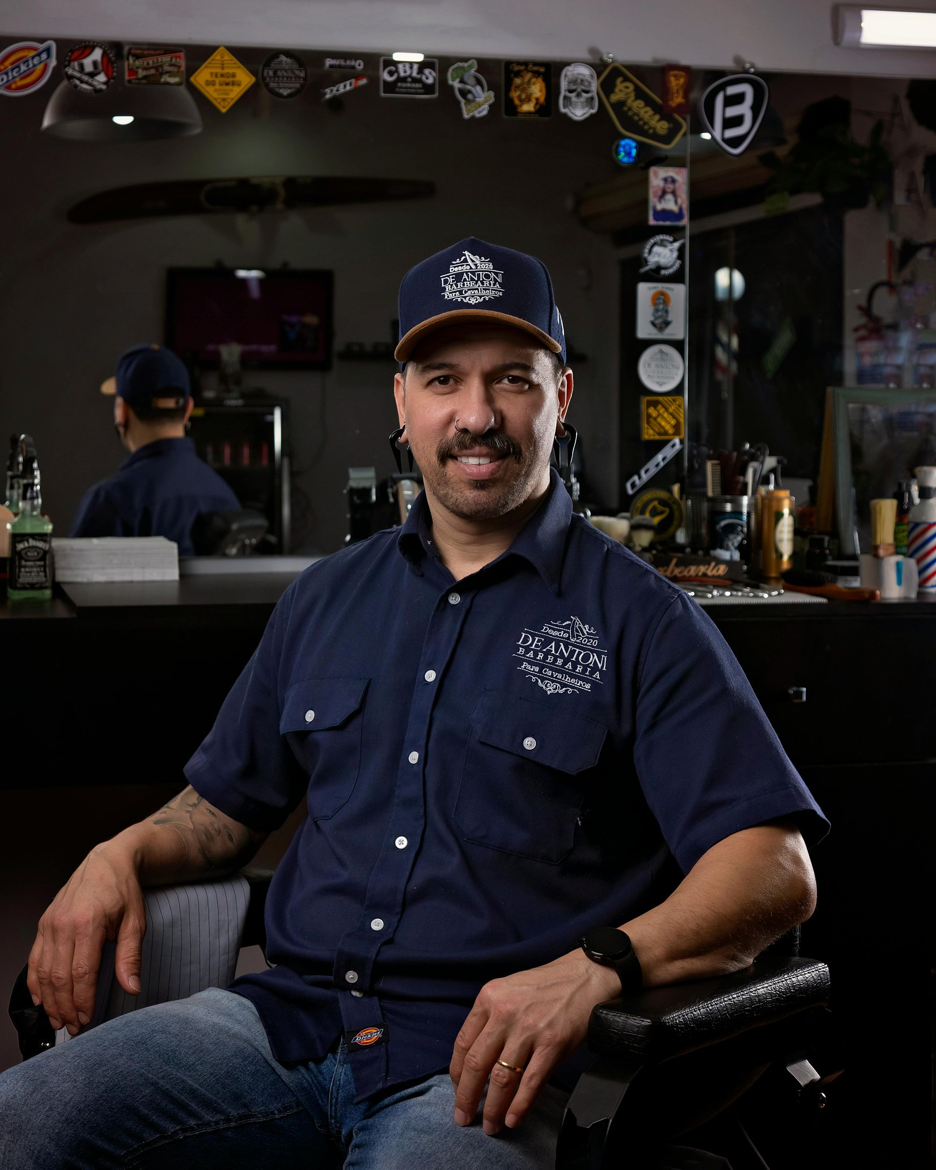 Man in a barber shop, smiling, wearing a blue work shirt and cap, sitting in a barber chair.
