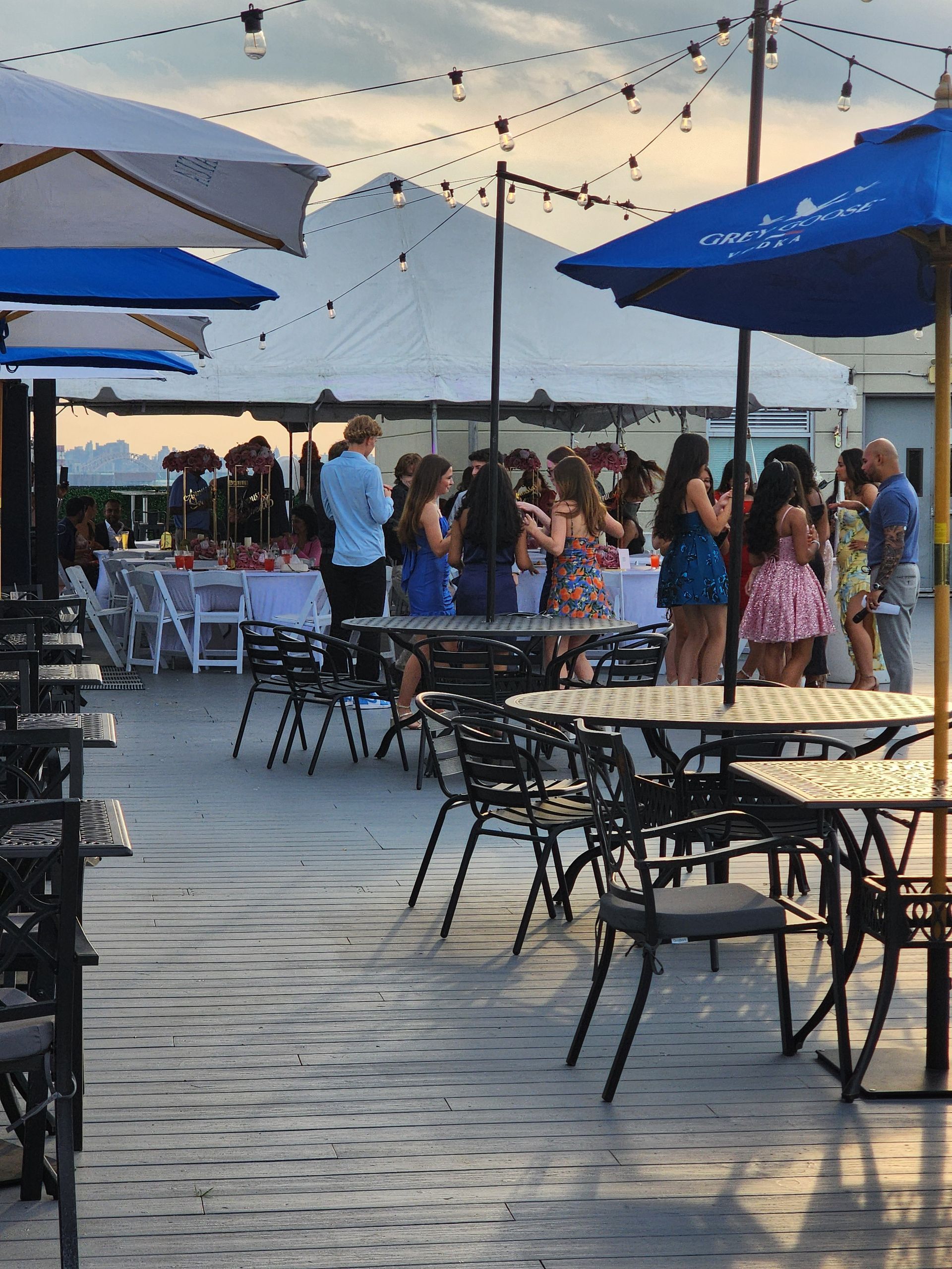 Outdoor event: people gathered near tables, tents, and umbrellas at dusk.