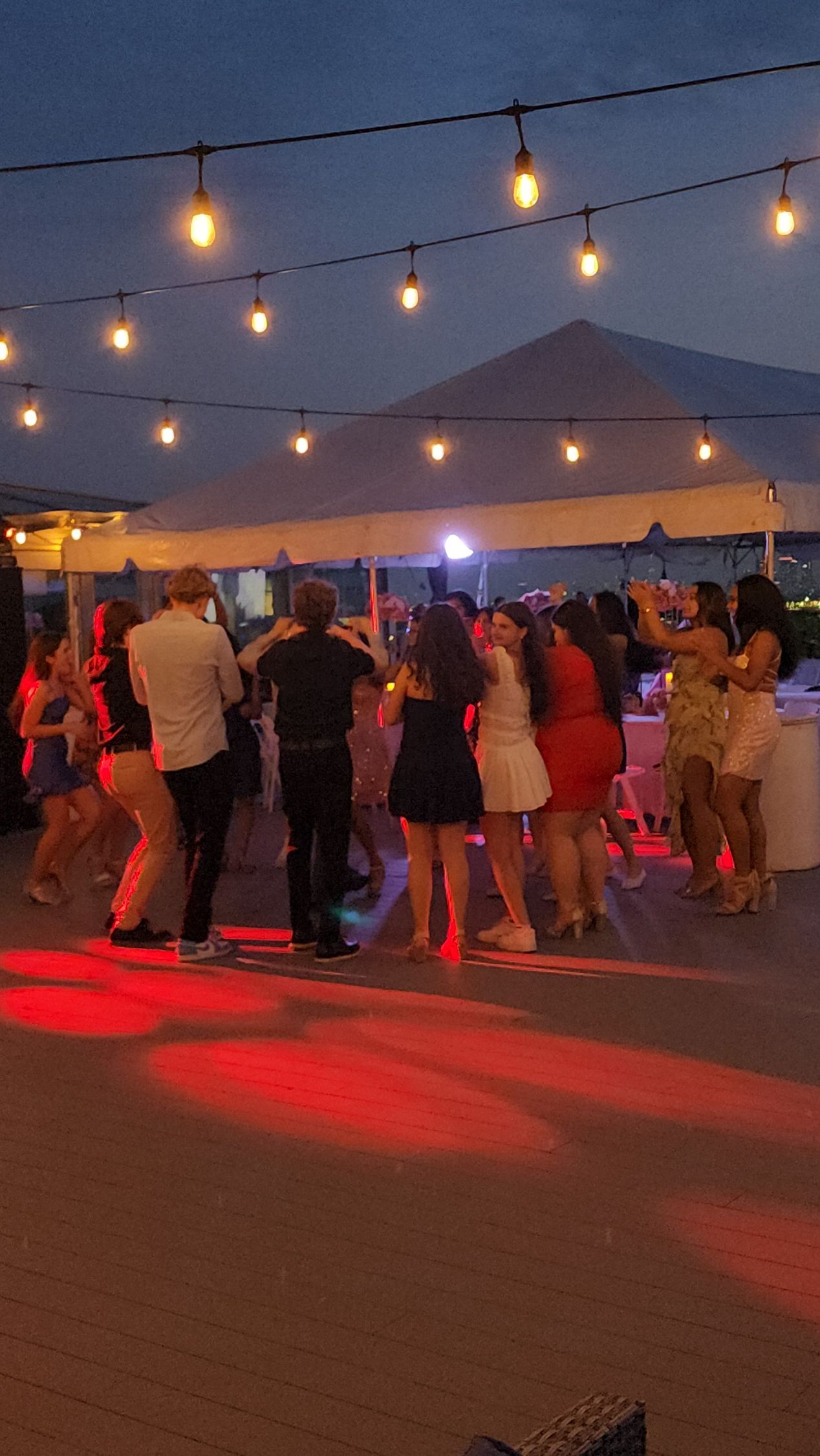 People dancing outdoors under string lights at night. A red glow illuminates the ground.