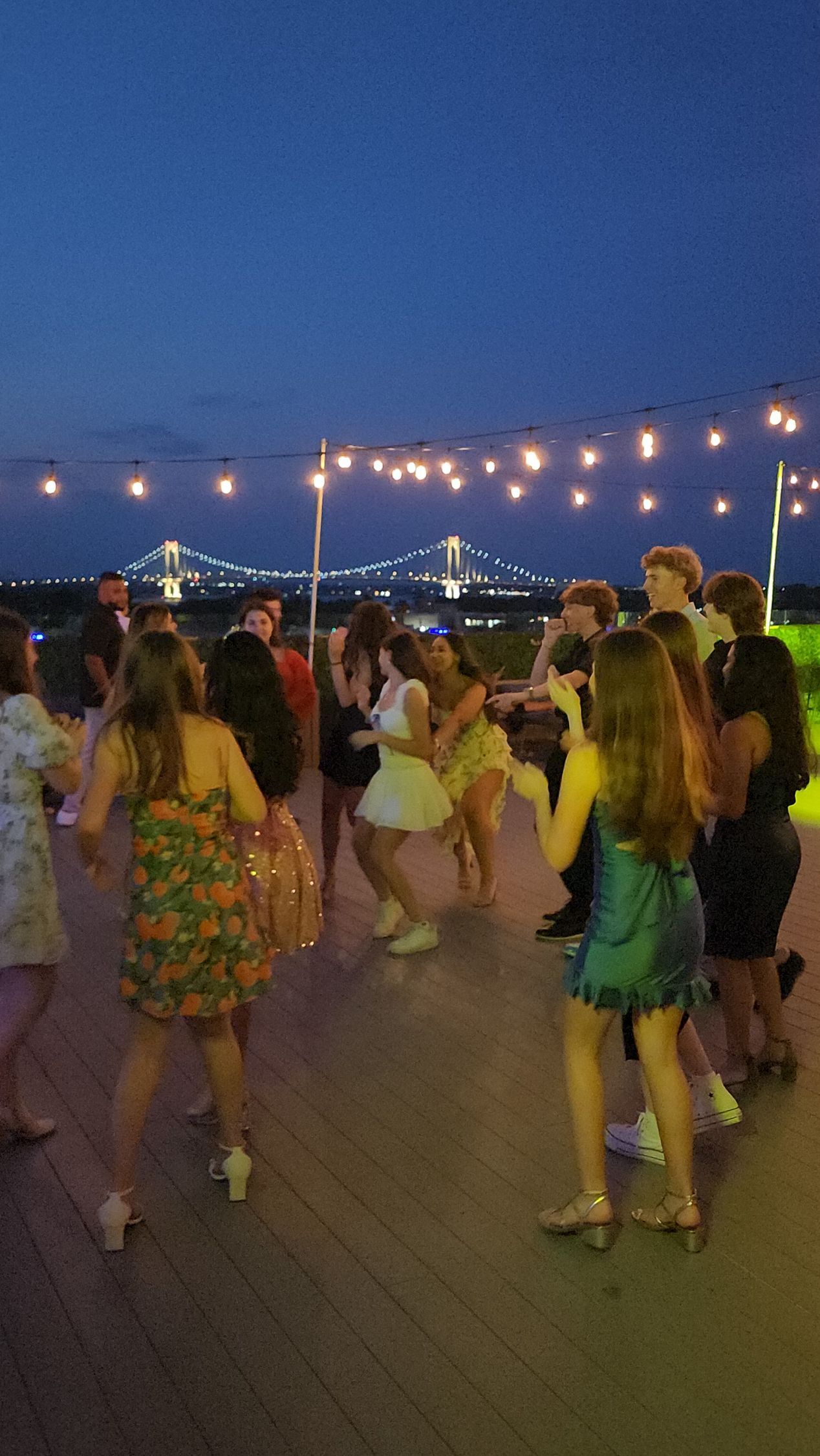 People dancing outdoors at night, string lights overhead, a bridge in the background.