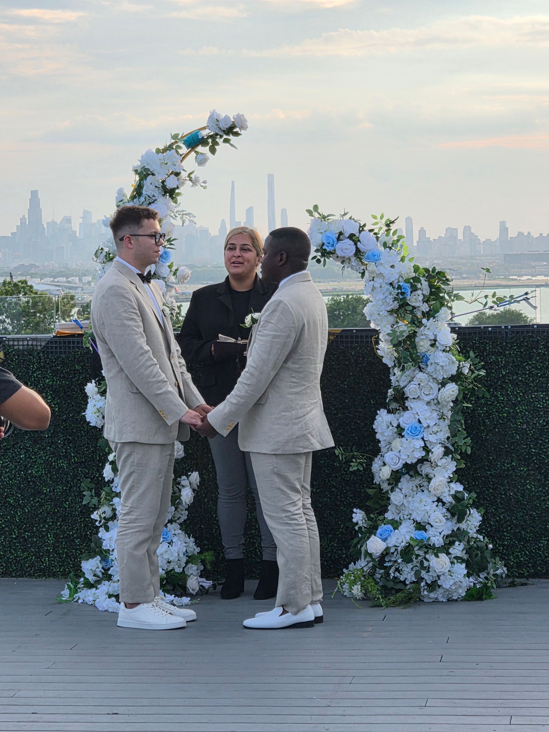 Two men in light suits holding hands under a white-and-blue floral arch at an outdoor rooftop wedding ceremony