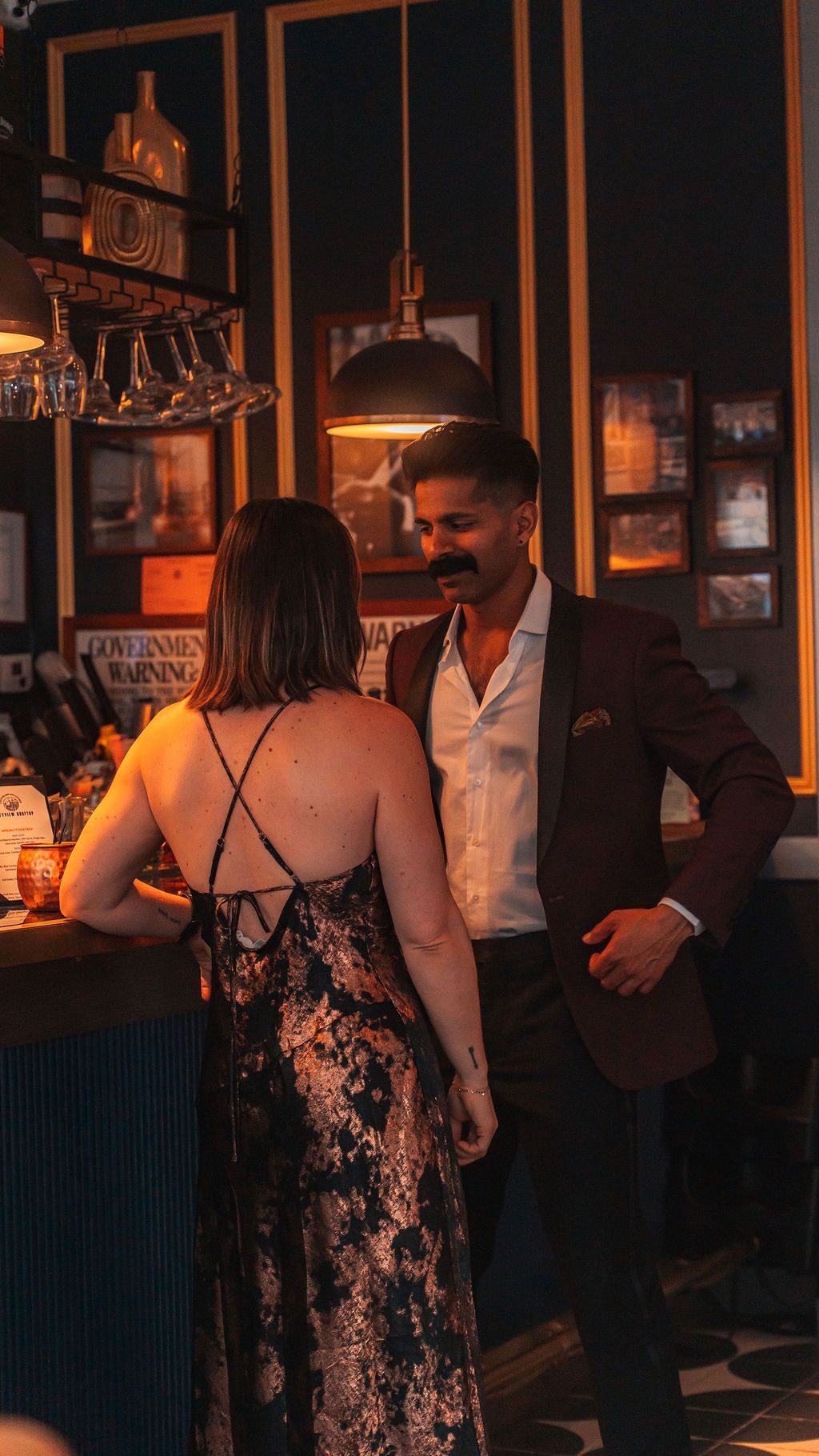 Couple at a bar, woman in a patterned dress, man in a suit jacket, warm lighting, leaning in close.