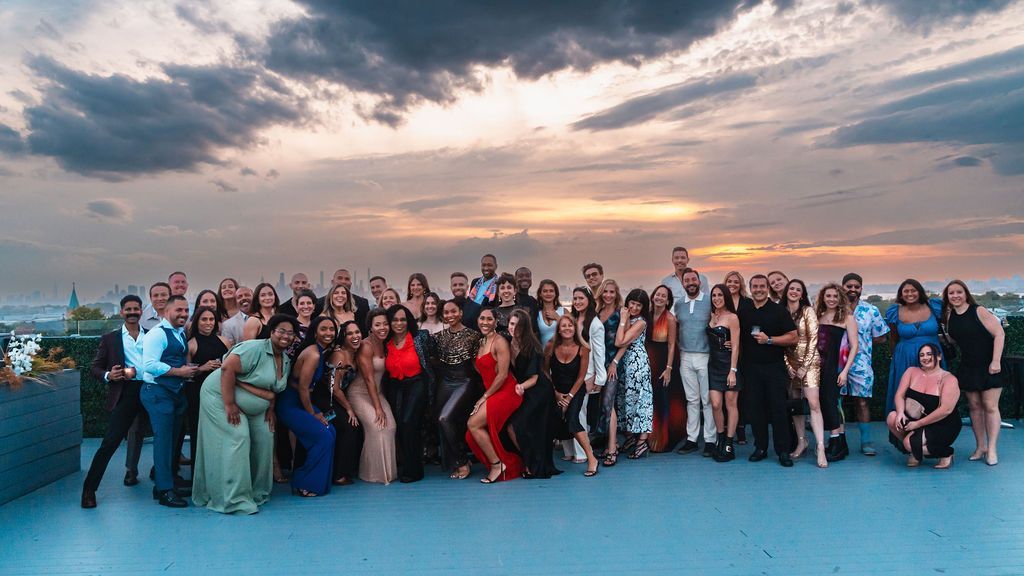 Group of people on a rooftop at sunset; diverse attire, smiling, and posing for a group photo.