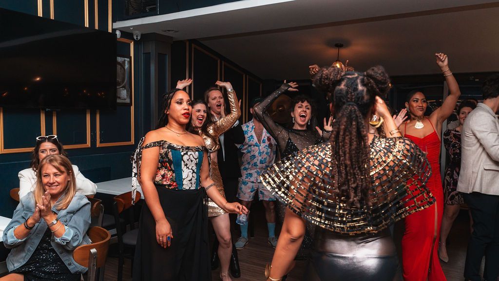 People dancing in a well-lit room with blue walls. Some wear formal attire, joyful expressions, raising arms.