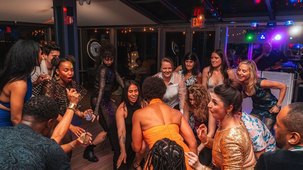 People dancing at a party in a well-lit space; some smiling, laughing, or singing, with lights in the background.