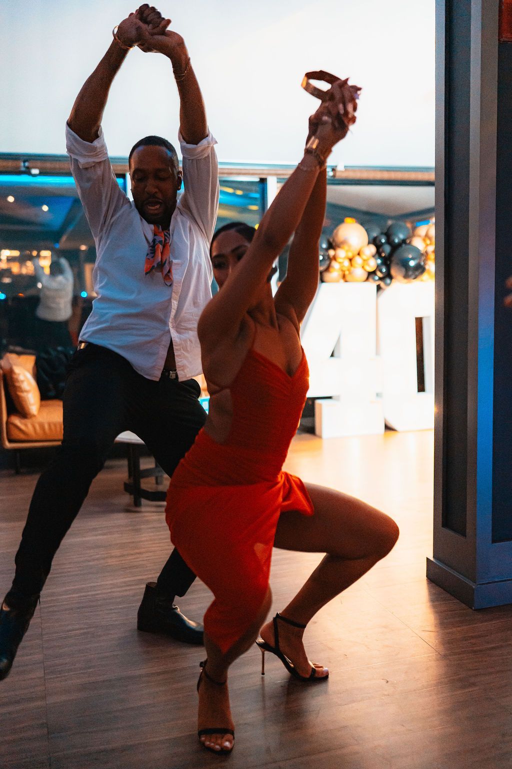 Couple dancing salsa, man in white shirt and woman in red dress with arms raised, on a wooden floor.