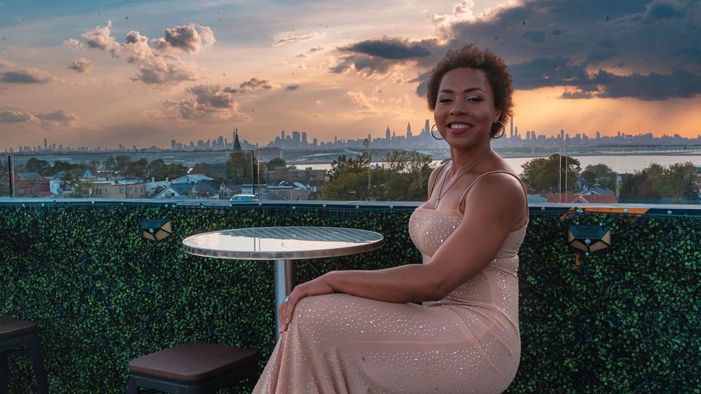 A woman in a long dress is sitting at a table with a view of the city.
