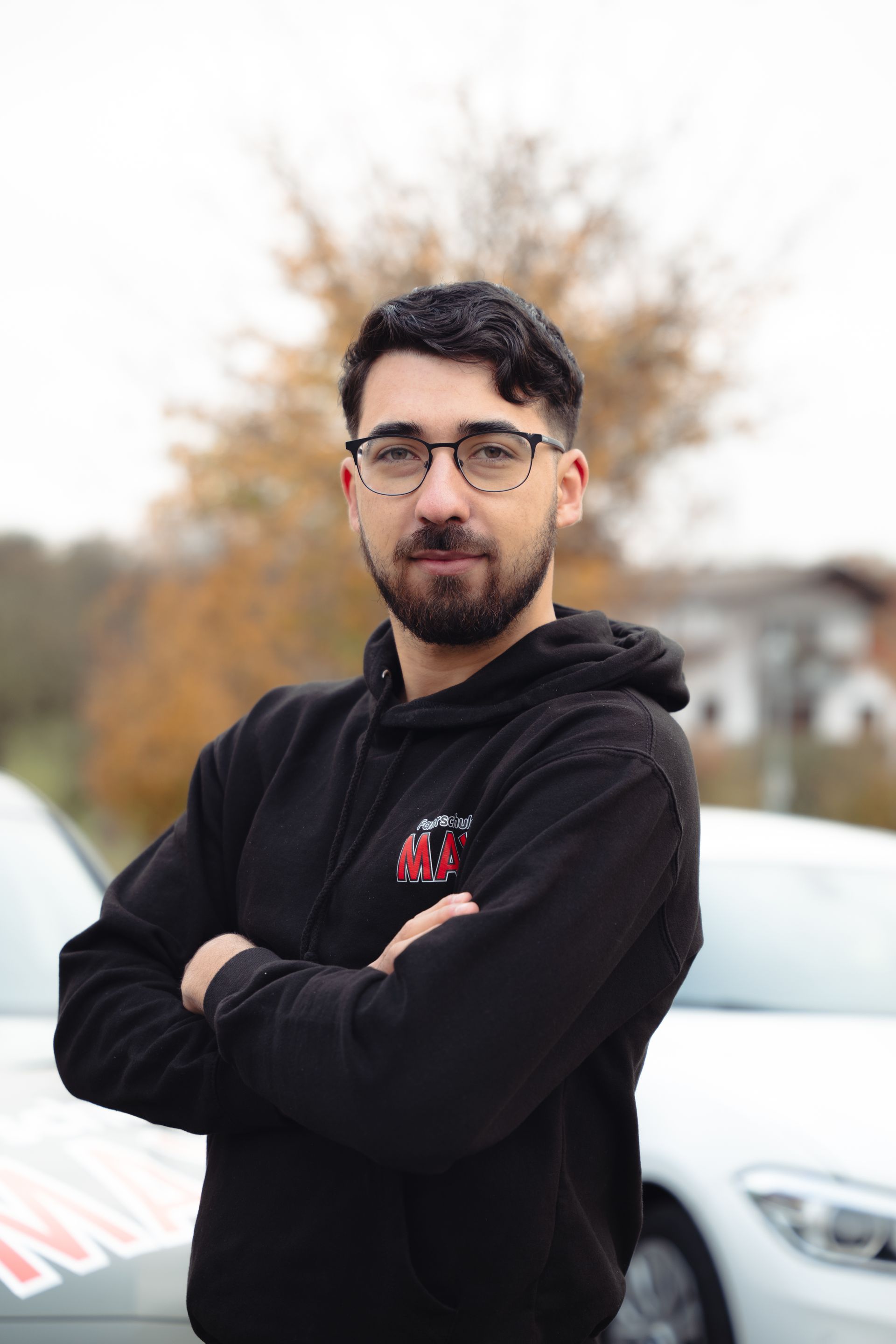 Ein Mann mit Bart und Brille steht mit verschränkten Armen vor einem Auto.