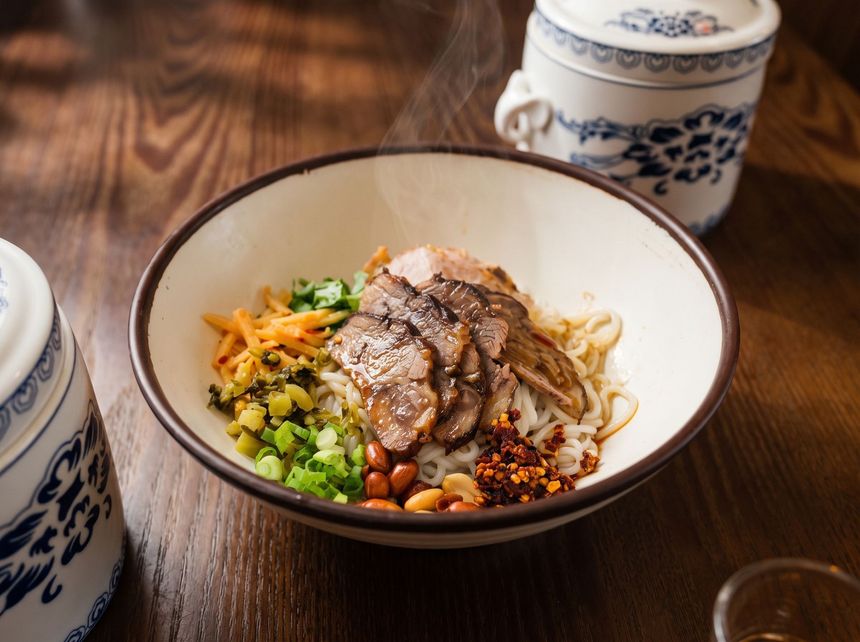Bowl of noodle soup with sliced meat, scallions, and chilies on a wooden table