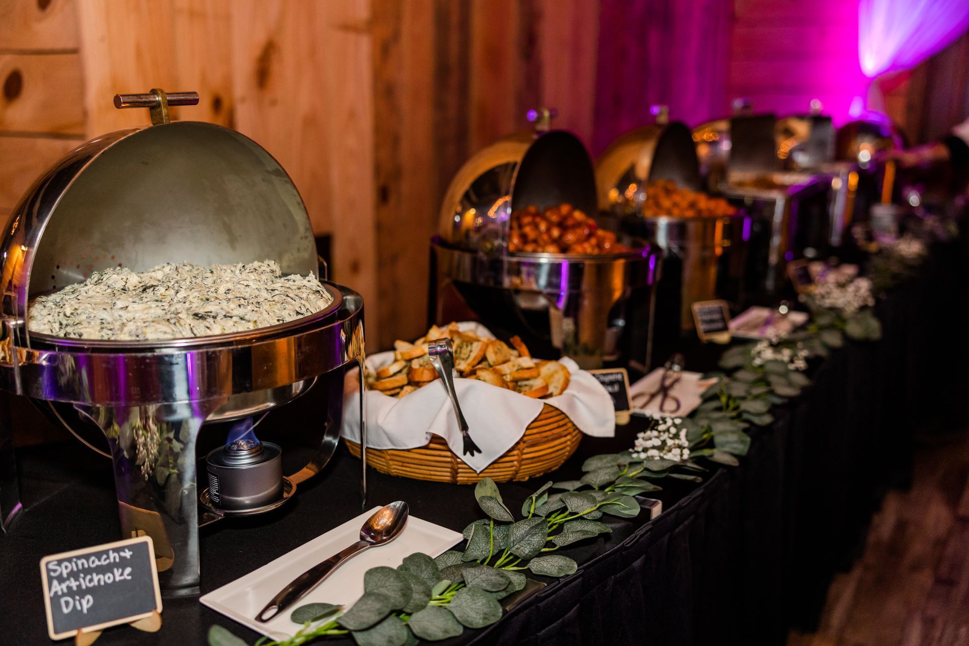 Buffet table with various food warmers and dishes, including a dip, rolls, and other entrees. The setting is indoors with wood paneling.
