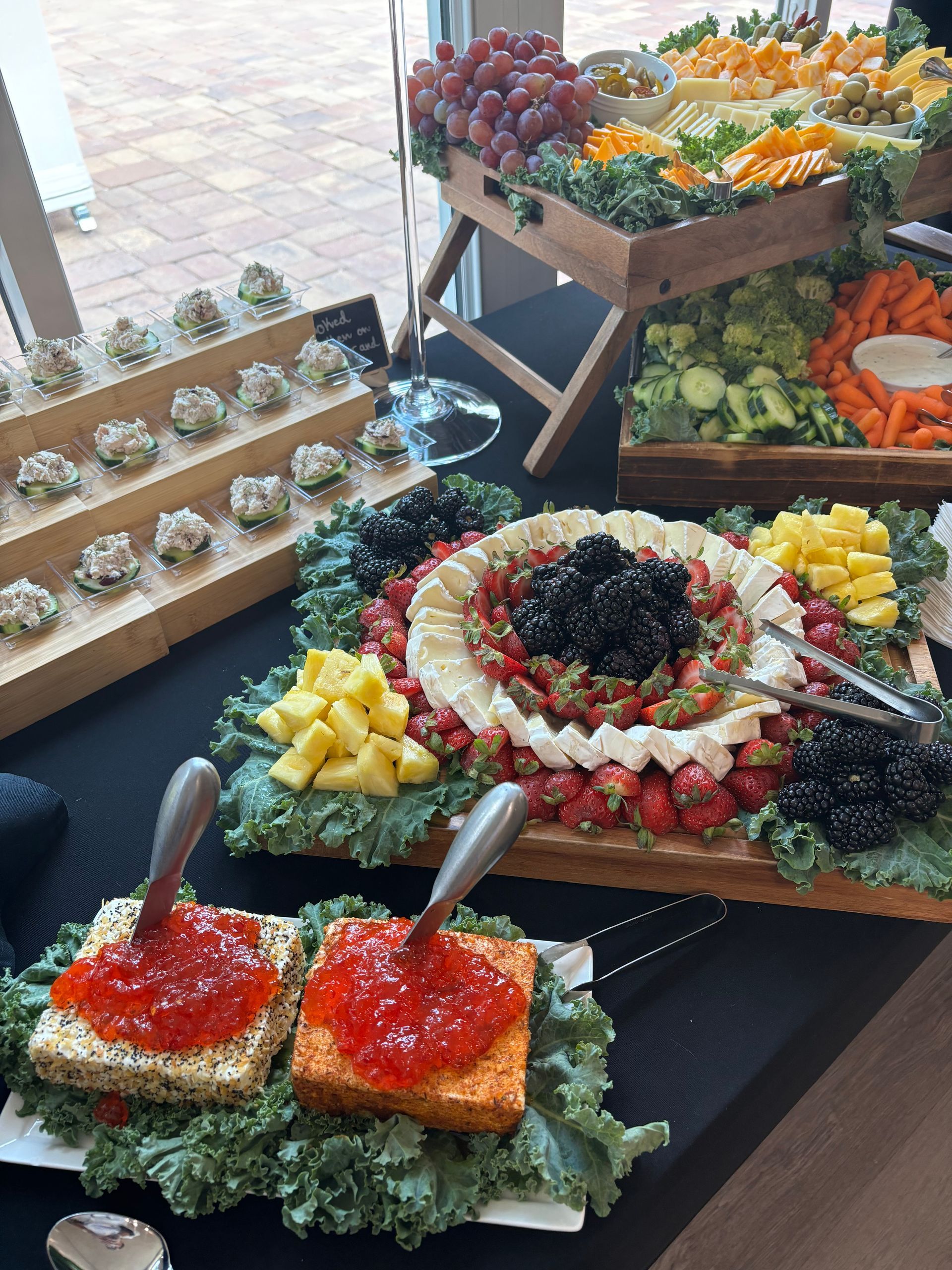 Buffet table with food displays: cheese, fruit, vegetables, and small appetizers, with a brick patio visible in the background.