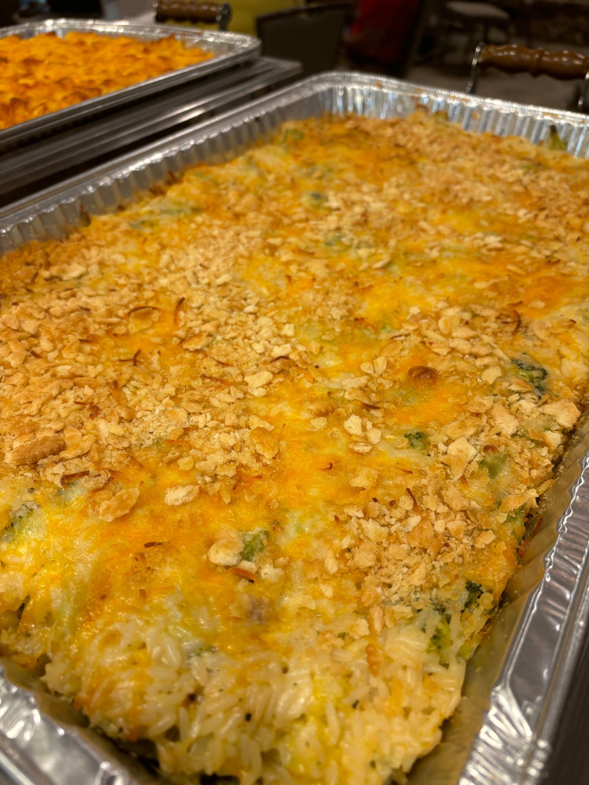 Overhead shot of a casserole in a foil pan, topped with melted cheese and breadcrumbs. Another pan is visible in the background.
