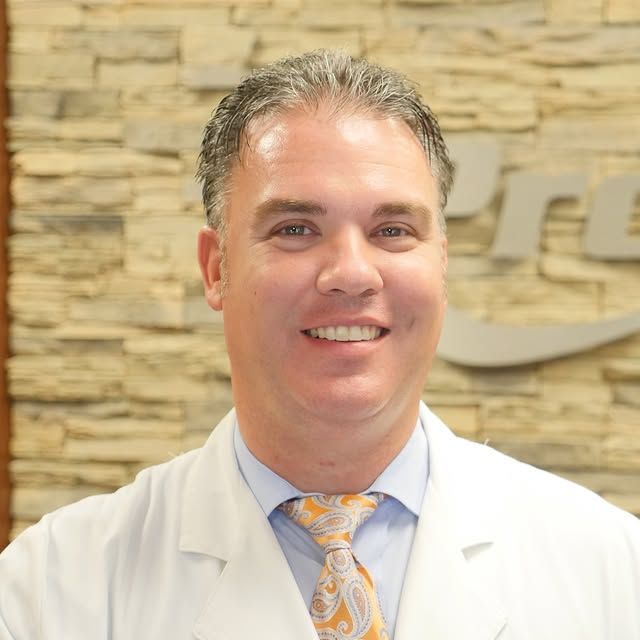 Smiling man in a white coat and patterned tie standing before a stone wall