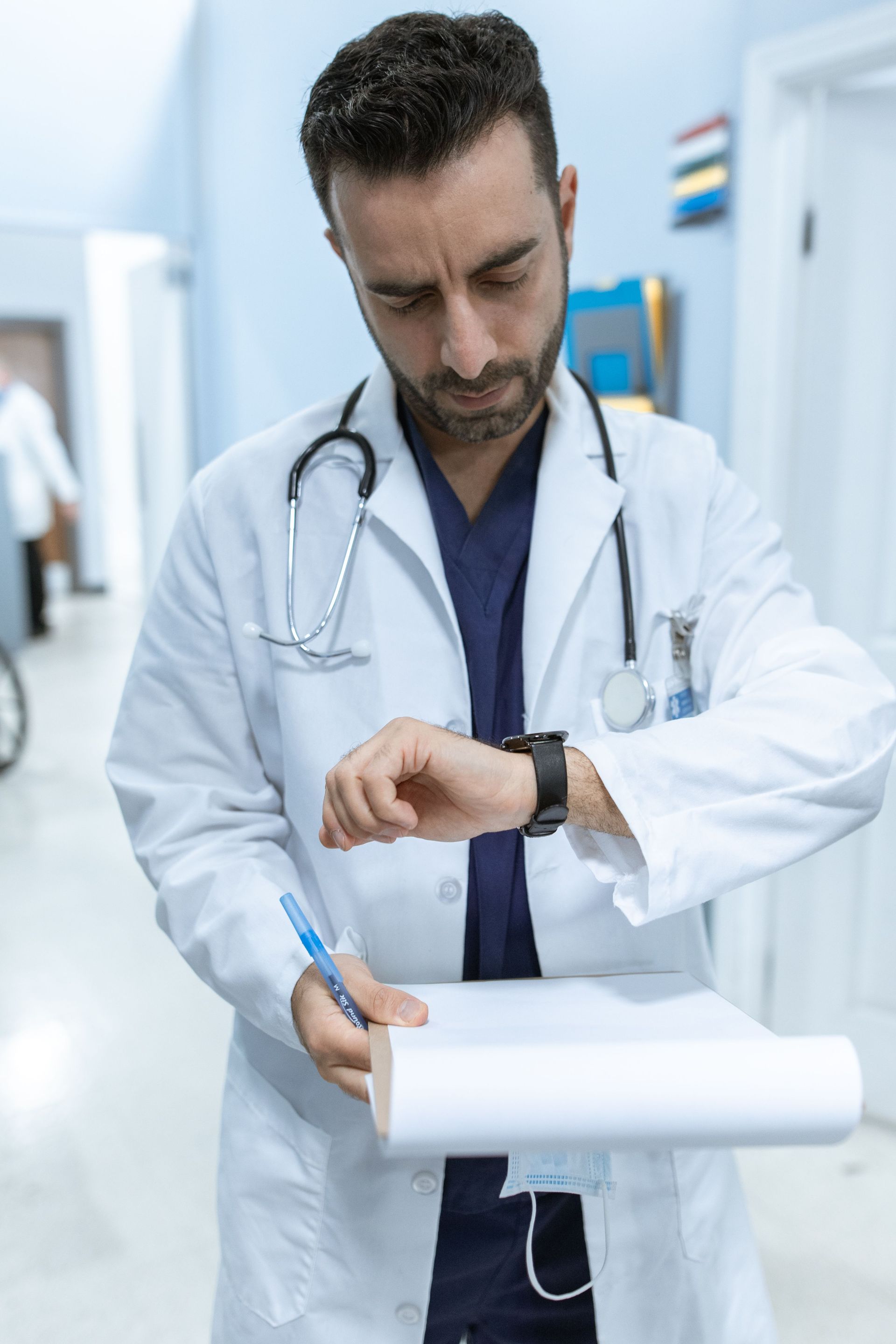 Doctor in a white coat checking his watch while holding a clipboard in a hospital corridor