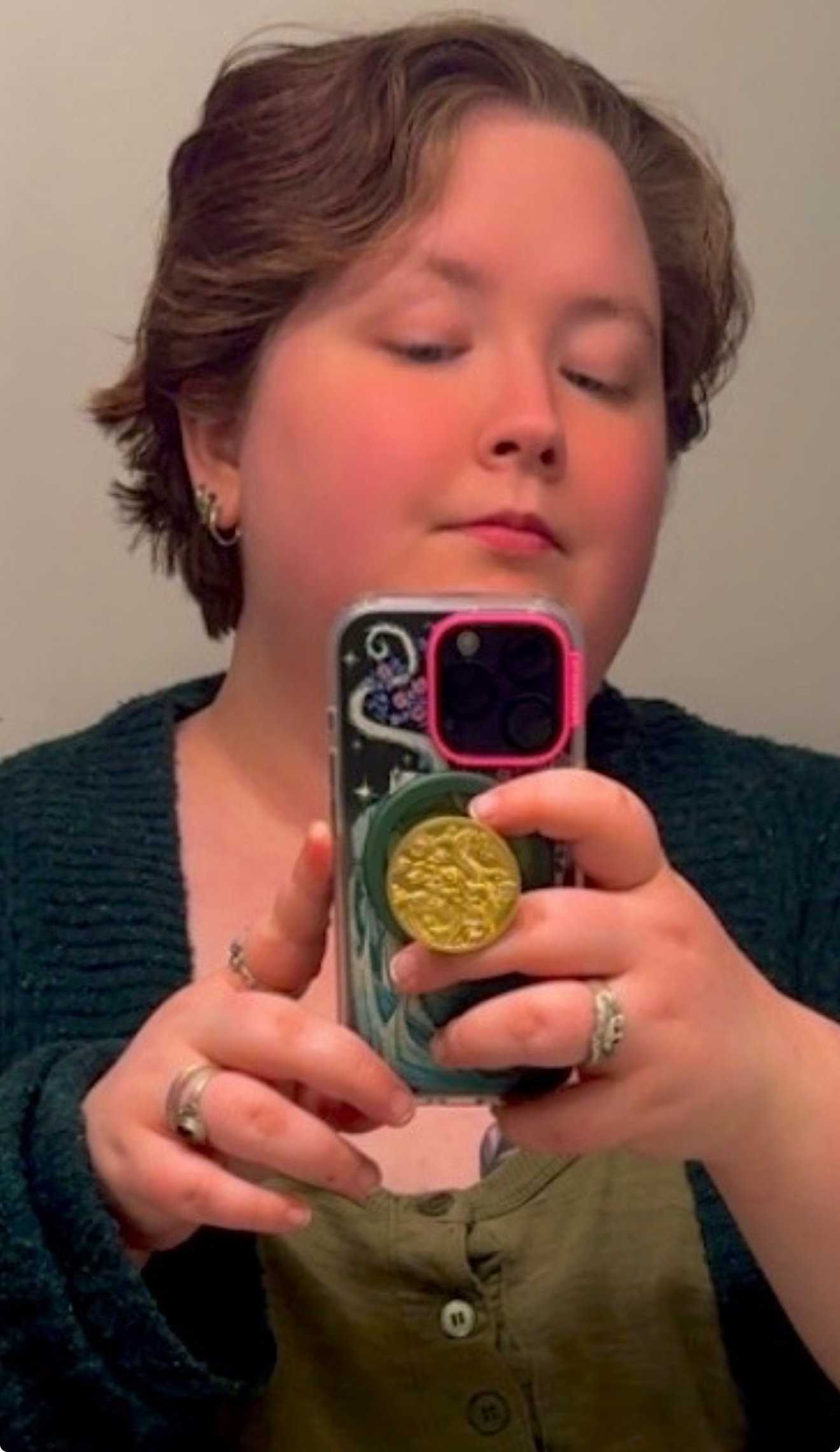 Person holding phone taking a selfie, wearing a green sweater and rings. Short brown hair.