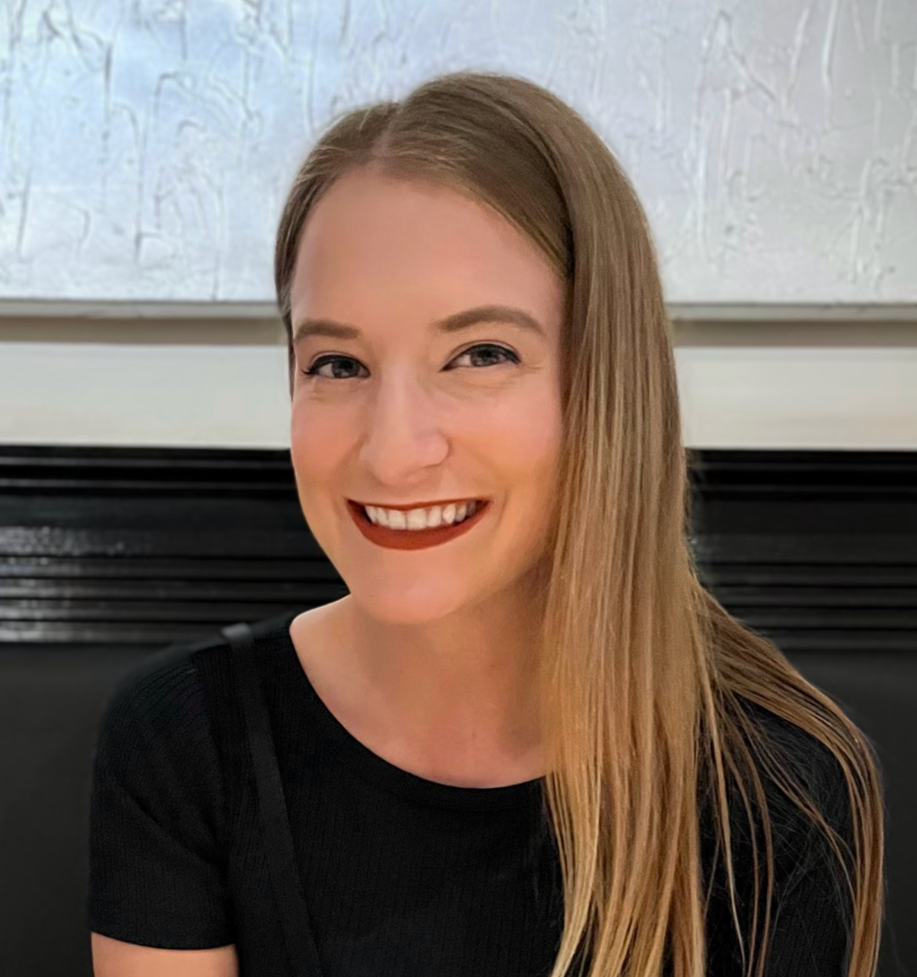 Woman with long blonde hair smiles, wearing a black top. Background: painting and dark fireplace.