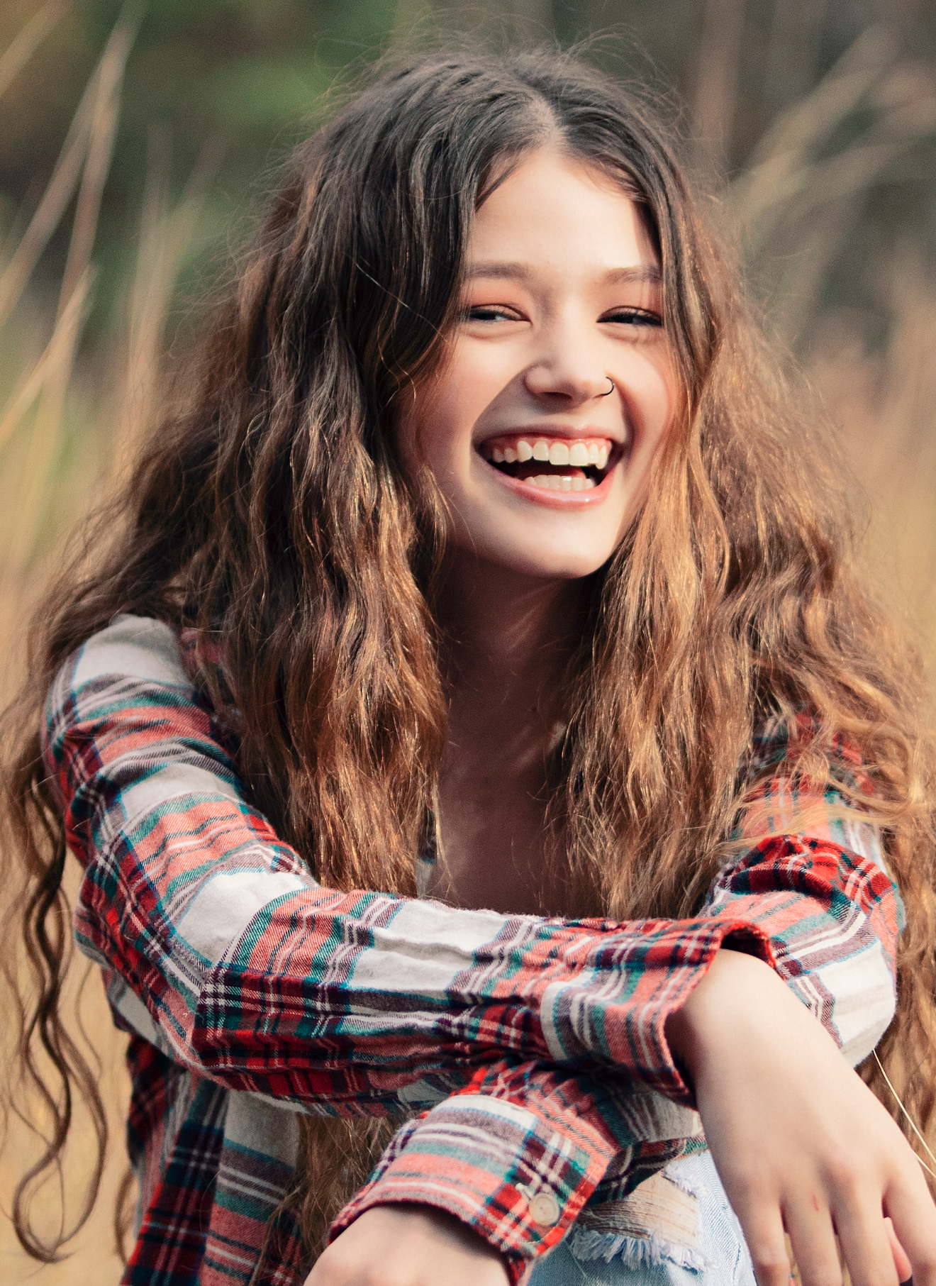 Woman with long, wavy brown hair, laughing outdoors in a plaid shirt.