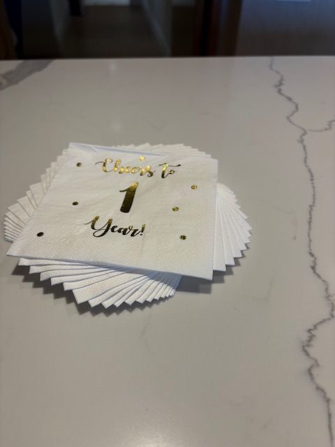 A stack of white cocktail napkins with gold writing that says Cheers To 1 Year.