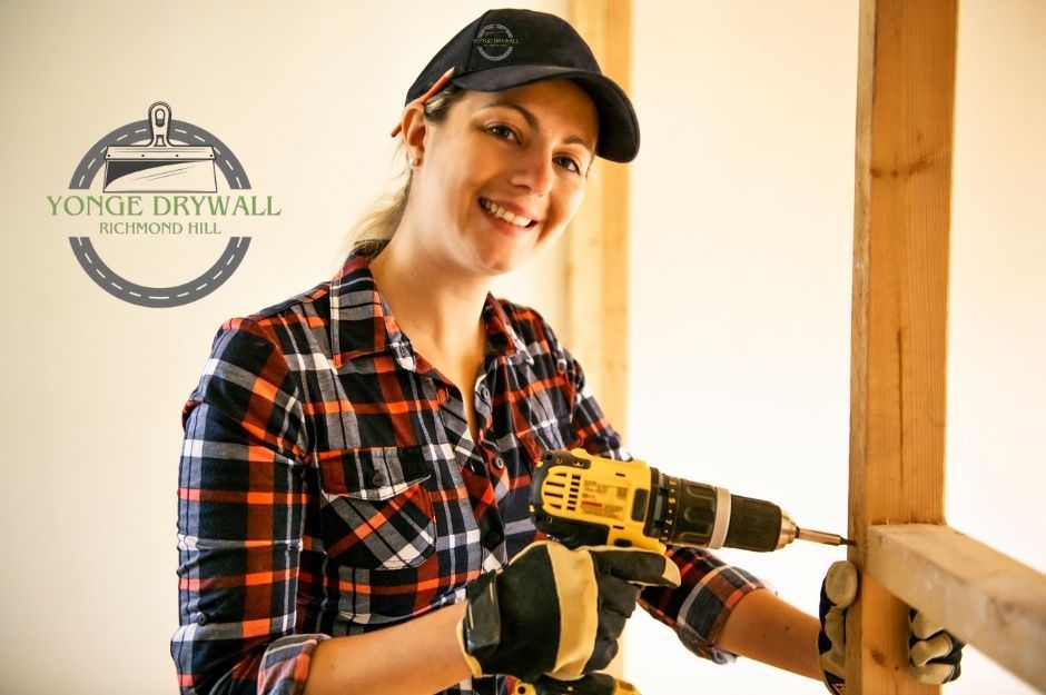 A drywall repair company worker, wearing a plaid shirt and black cap, uses a power drill on a residential construction project. She wears work gloves and smiles while working on wooden framing in a renovation space with white drywall walls near Ketelbey Ct, and Walkers Line, Burlington, ON.