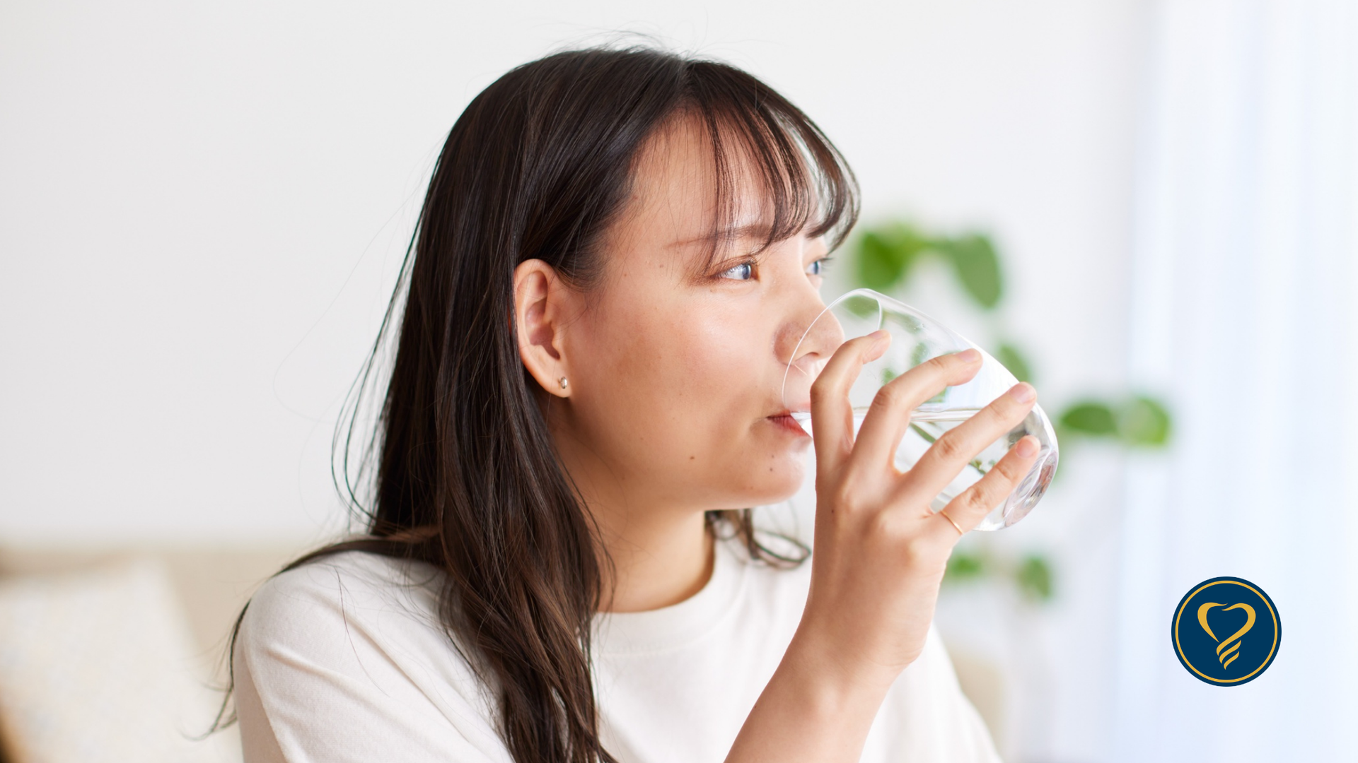 A person in a white shirt drinks from a clear glass of water in a brightly lit, minimalist indoor setting.