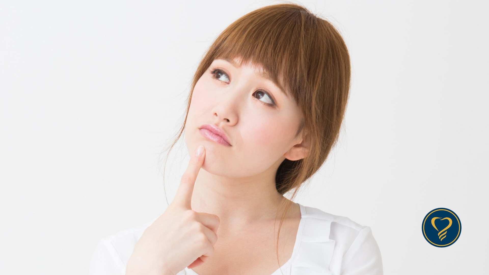 A person with brown hair looks upward with a thoughtful expression, touching their chin with a finger against a white wall.
