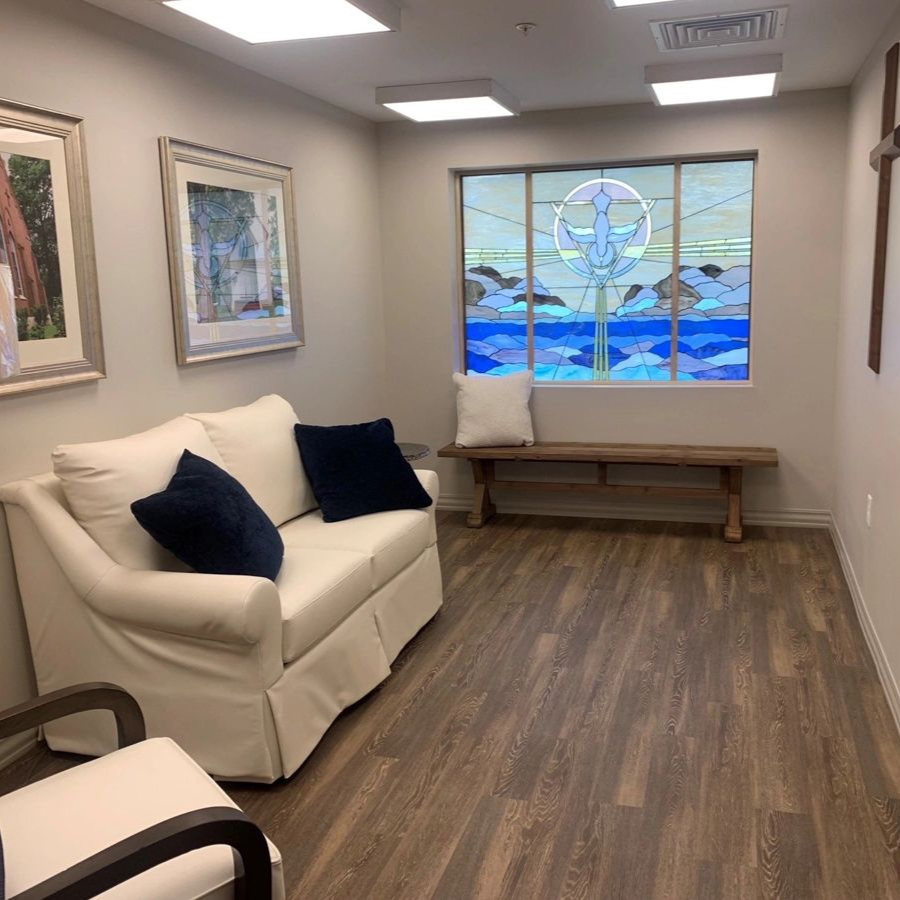 Waiting room with stained glass window, white couch and two navy pillows, bench and wood floors.