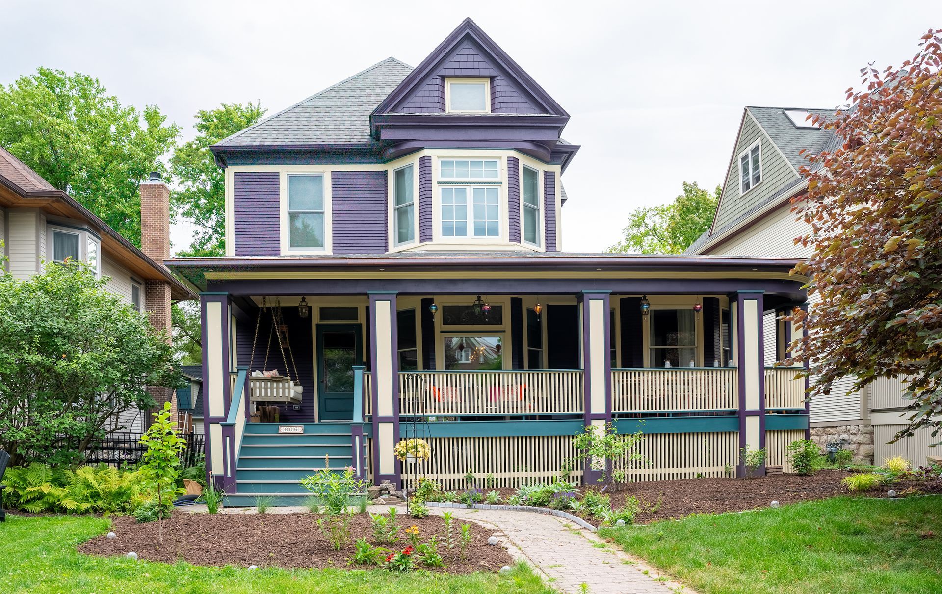 A large purple house with a large porch is sitting on top of a lush green lawn.