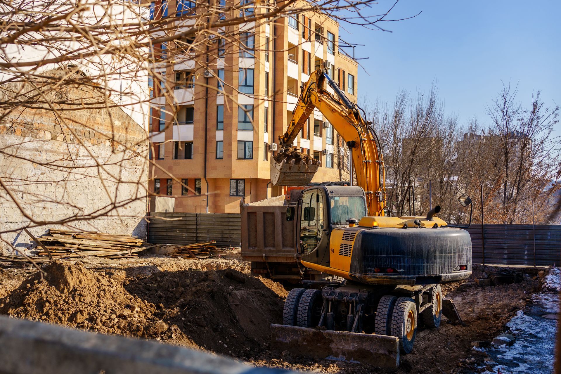 Excavator digging beside a tan apartment building on a dirt construction site in winter