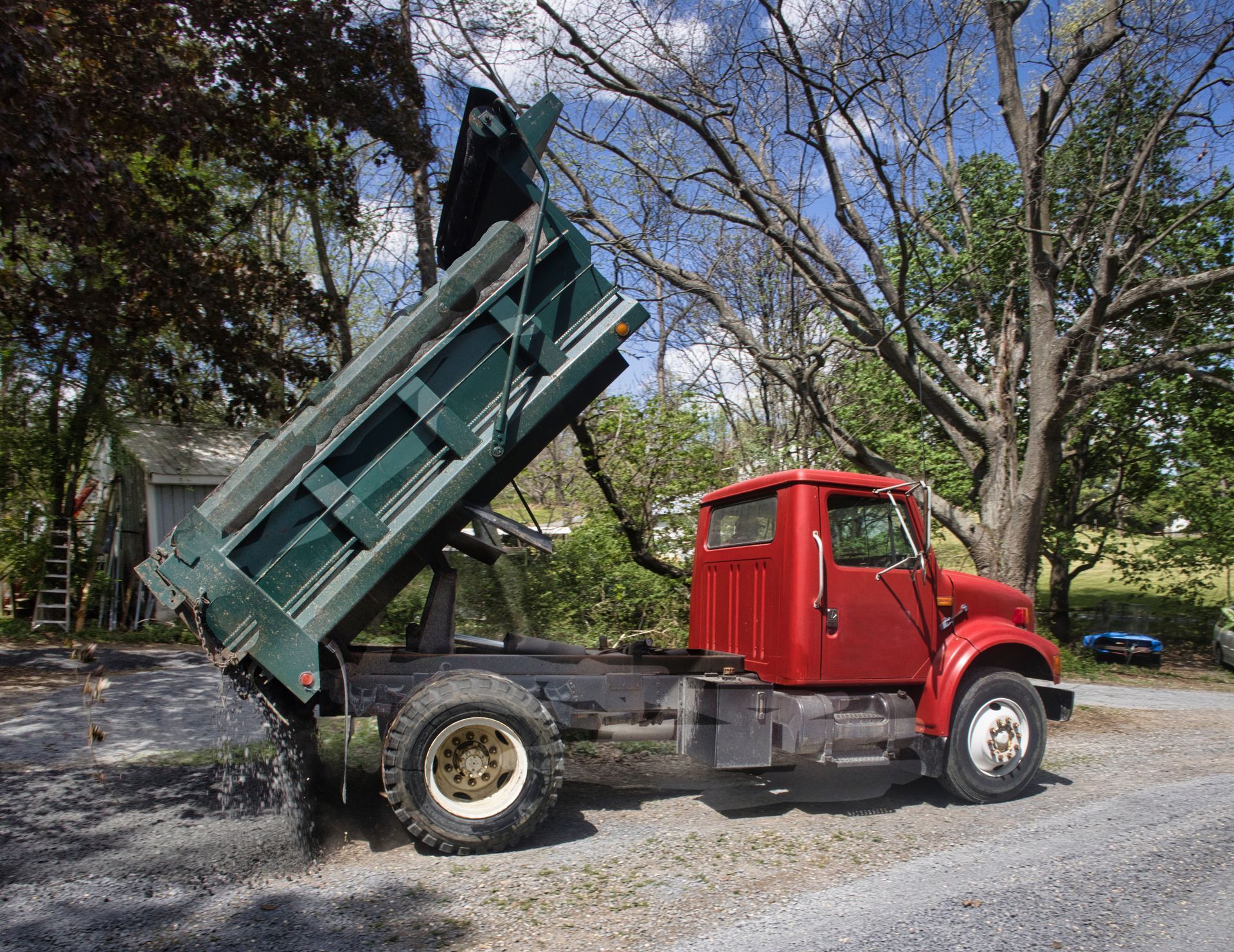 Red dump truck with raised green bed parked on a gravel road beside trees Red dump truck with raised green bed parked on a gravel road beside trees