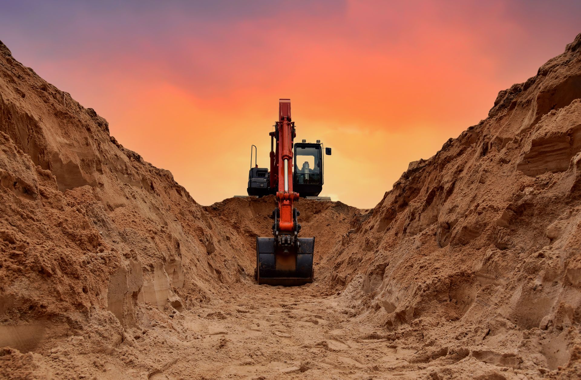 Excavator digging in a sandy trench at sunset under an orange sky