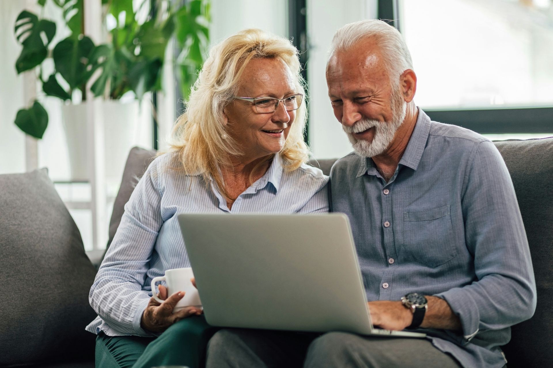 An older couple smiles, looking at a laptop together on a couch. The woman holds a mug.
